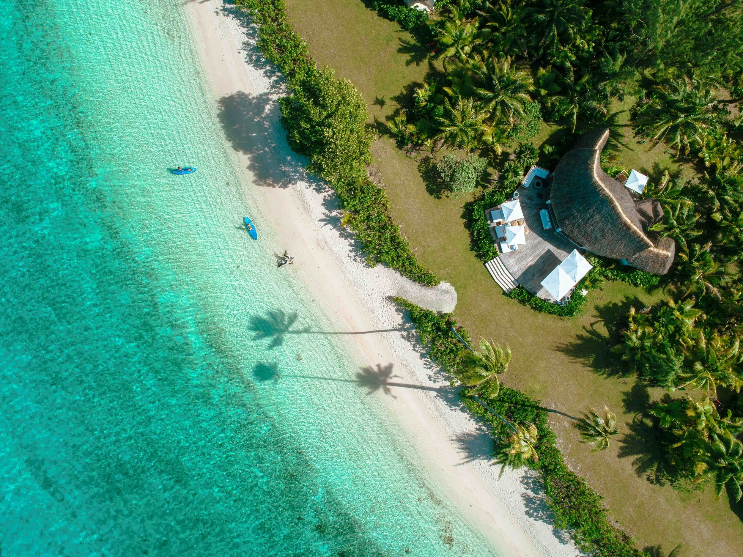 Vue aérienne d'une plage de sable blanc avec trois kayaks dans l'eau turquoise, entre des palmiers et un bungalow avec terrasse équipé d'ombres et de mobilier.