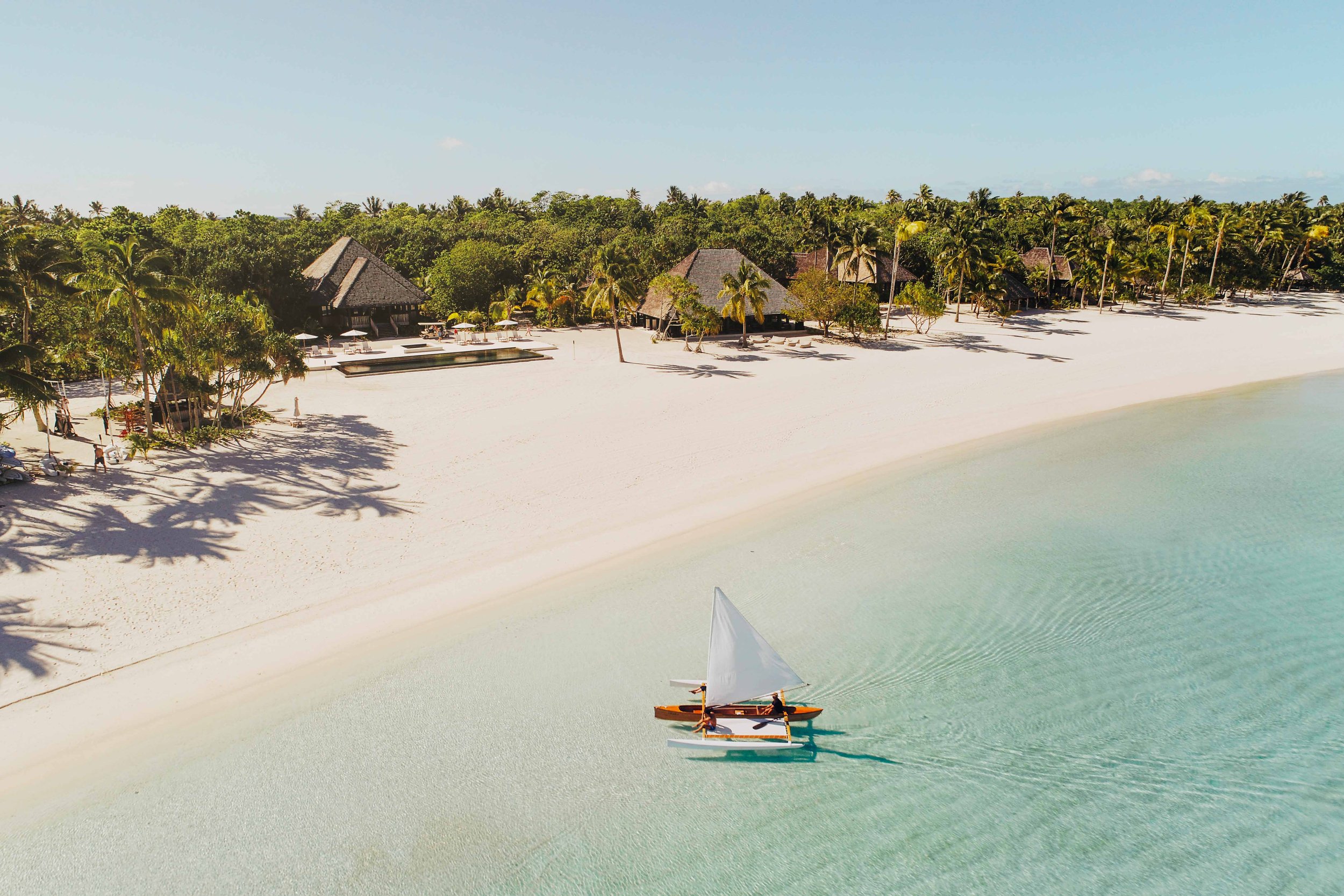 Plage de sable blanc avec un bateau à voiles sur l'eau turquoise et des maisons en huttes entourées de palmiers en arrière-plan.
