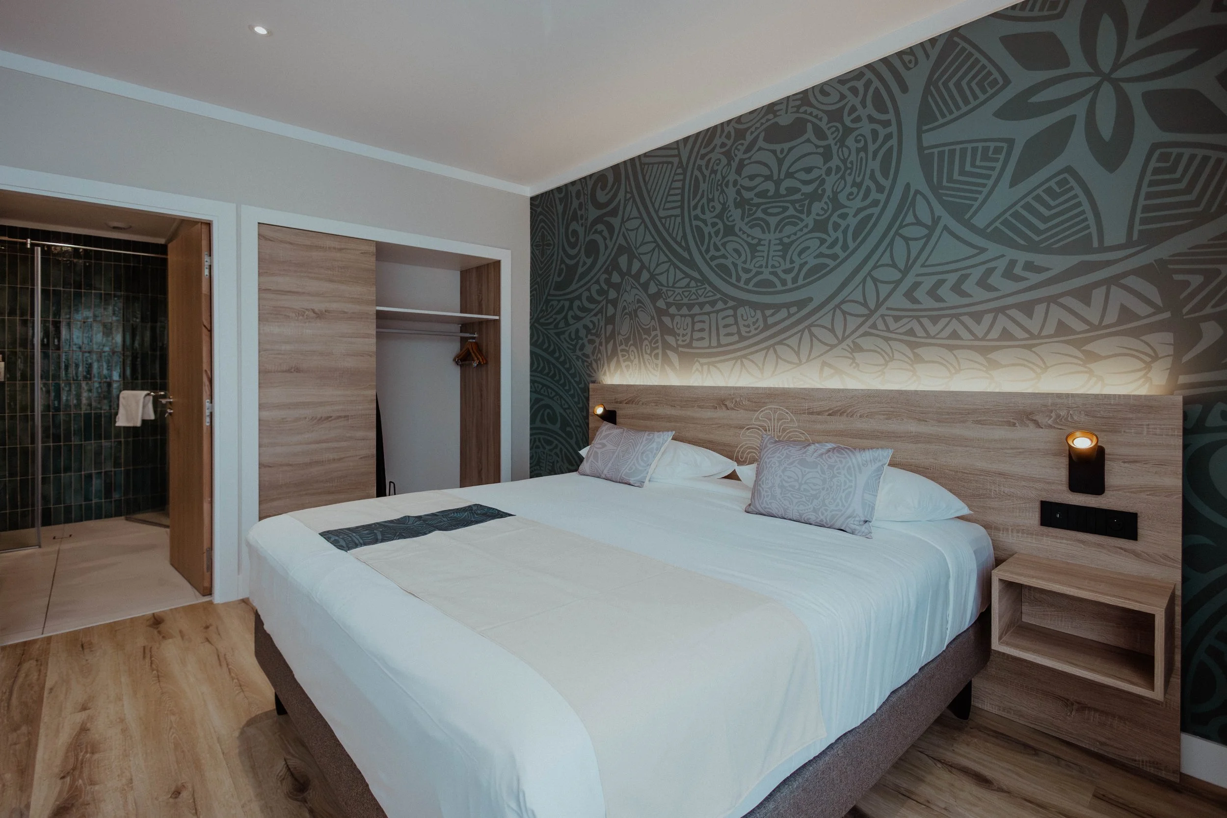 Chambre moderne avec lit double, deux oreillers, tête de lit en bois, mur décoratif avec motif tribal, lampes de lecture, et salle de bain ouverte avec douche à gauche.