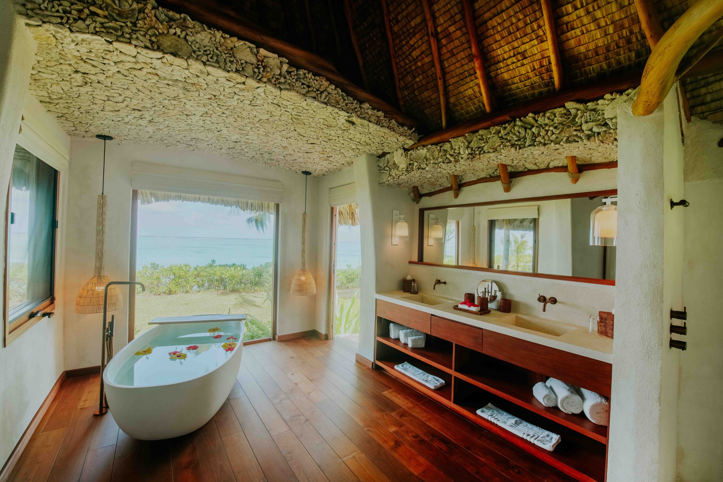 Salle de bain avec une baignoire en vue, située près d'une baie vitrée offrant une vue sur la mer et la nature, avec un lavabo double, un miroir, et une décoration naturelle et chaleureuse.