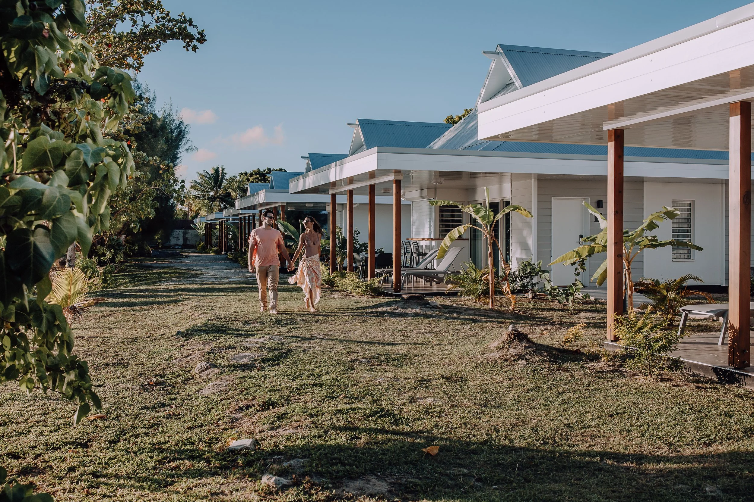 Un couple se promène main dans la main devant des villas modernes avec porches, entourées de plantes tropicales, par une journée ensoleillée.