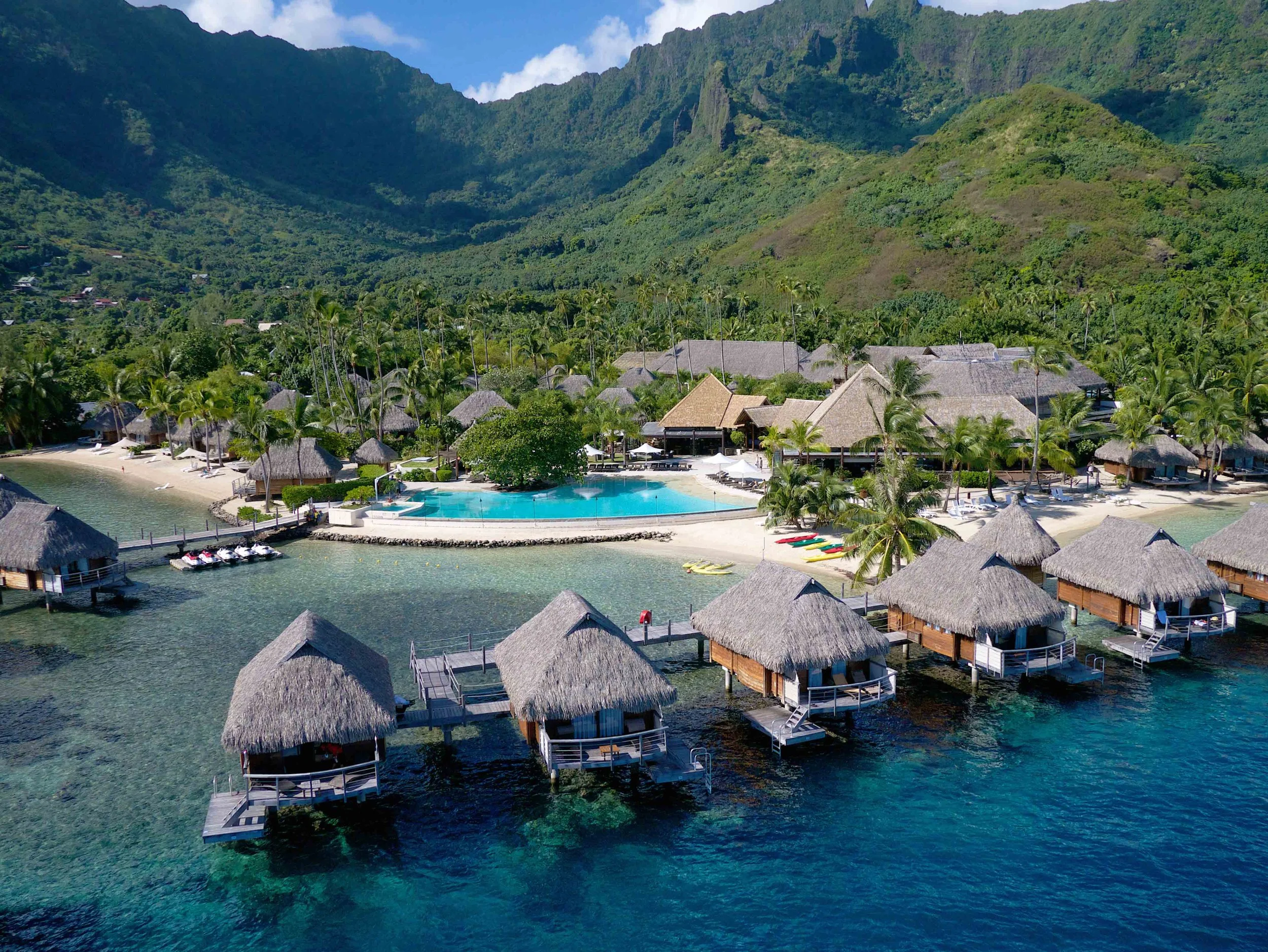 Complexe hôtelier avec villas sur pilotis, piscines et plages en bord de mer, entouré de végétation tropicale et de montagnes dans un paysage insulaire