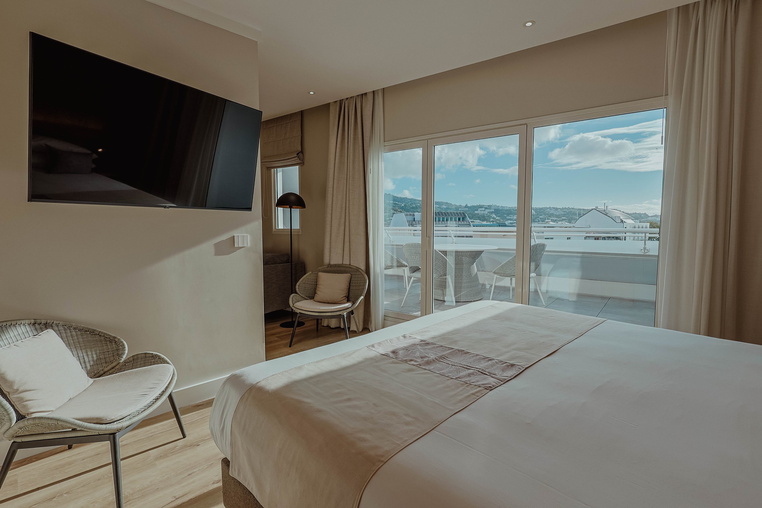 Chambre d'hôtel avec lit, fauteuils, télévision montée sur le mur, porte-fenêtre donnant sur un balcon avec vue sur la ville et le ciel ensoleillé.