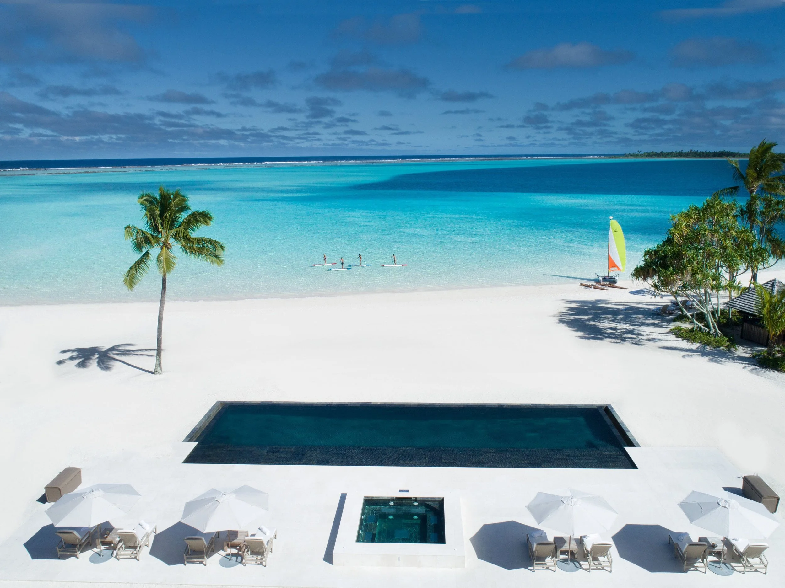 Plage de sable blanc avec des parasols et des chaises longues, une piscine à débordement et la mer turquoise en arrière-plan, avec un ciel partiellement nuageux.