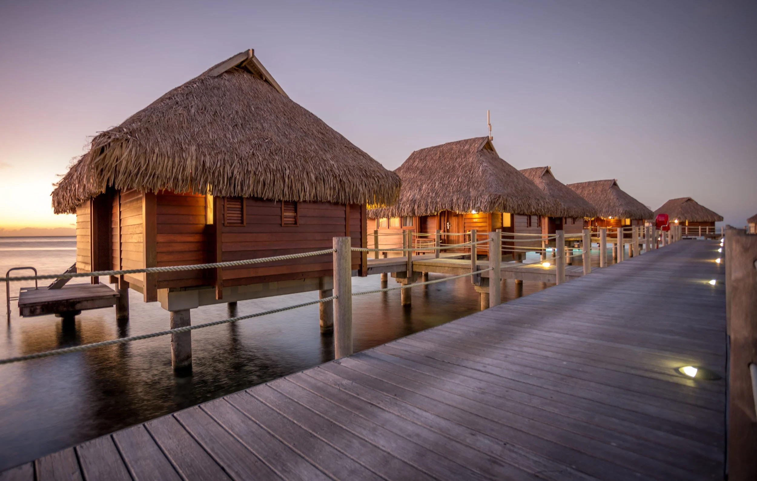 Bungalows en bois avec toits en chaume sur pilotis au dessus de l'eau, près d'une jetée en bois éclairée la nuit.
