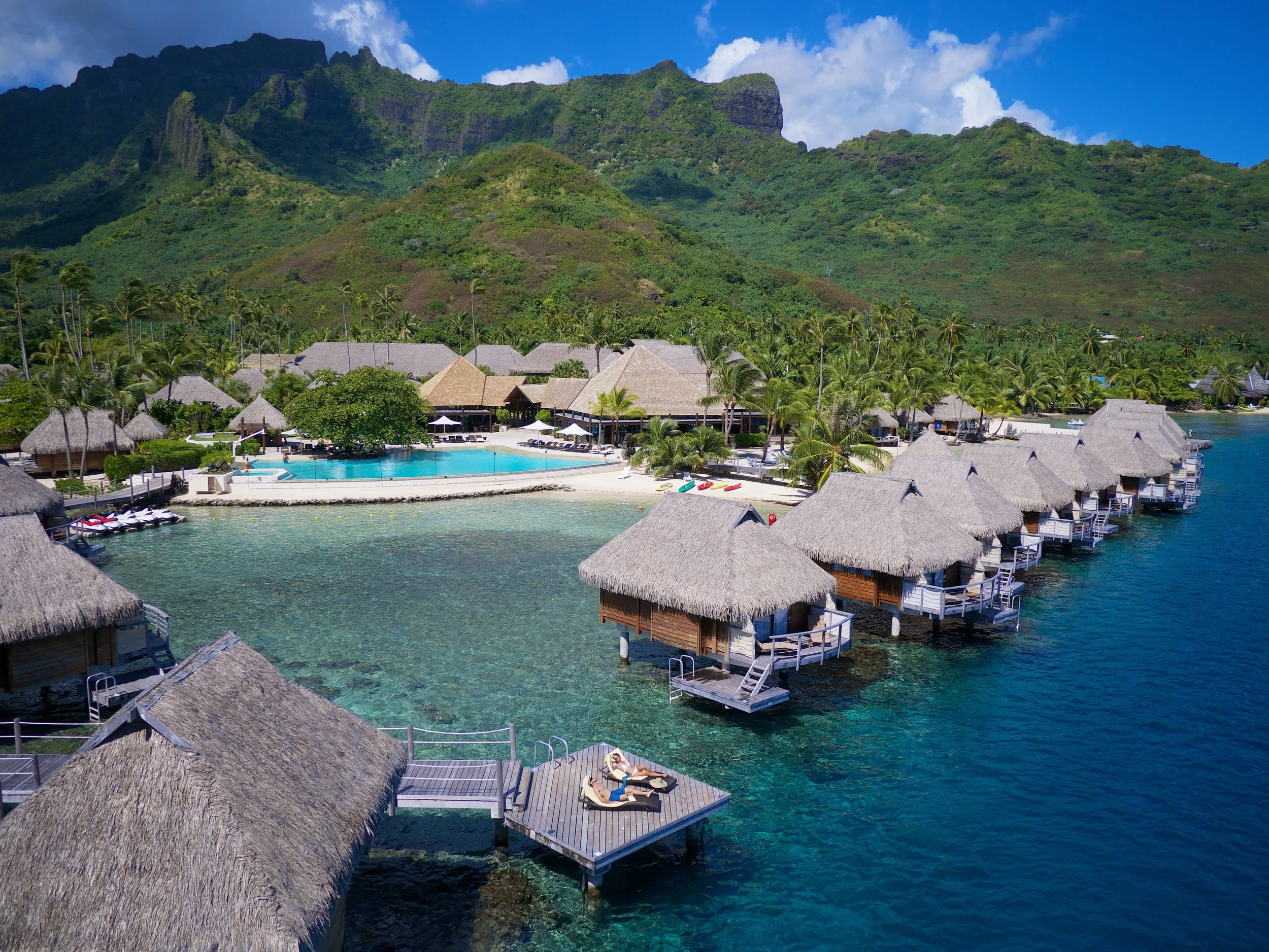 Bungalows overwater avec toits en chaume, une piscine et un paysage montagneux verdoyant en arrière-plan, dans une station balnéaire tropicale.