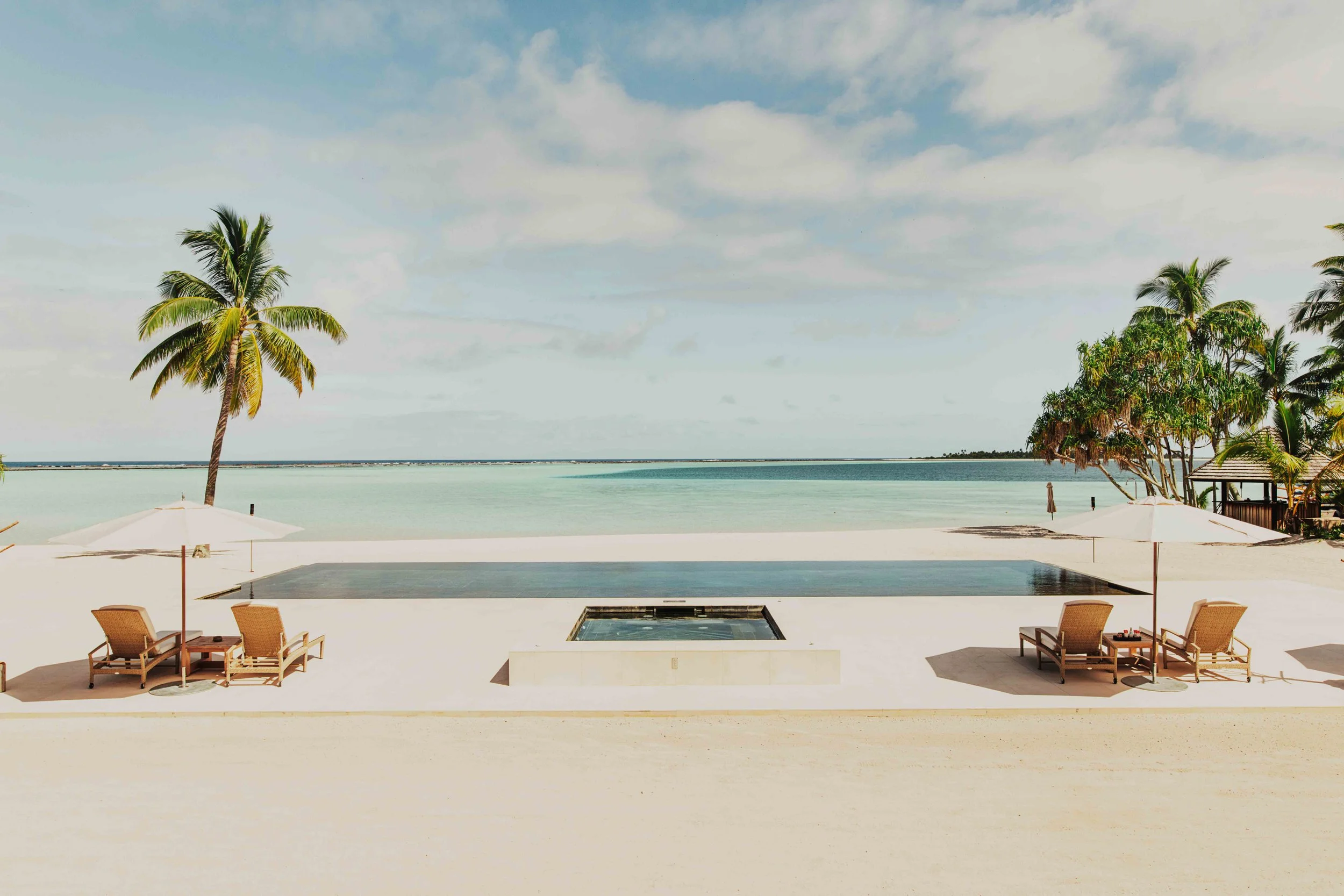 Plage de sable blanc avec des chaises longues, parasols, piscine, palmiers et vue sur l'océan calmes avec un ciel partiellement nuageux.