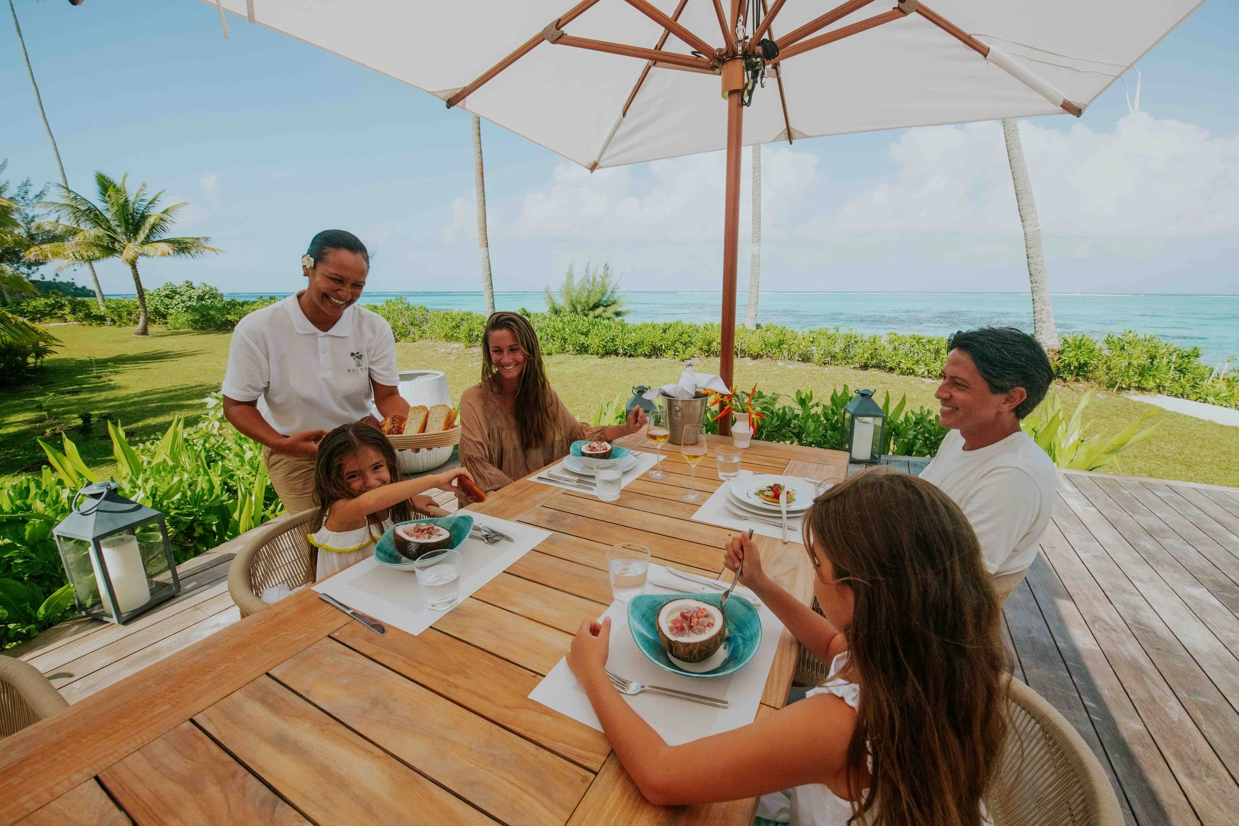 Groupe de cinq personnes dégustant un repas en plein air près de la mer, avec un parasol blanc, entouré de végétation tropicale, sous un ciel ensoleillé.