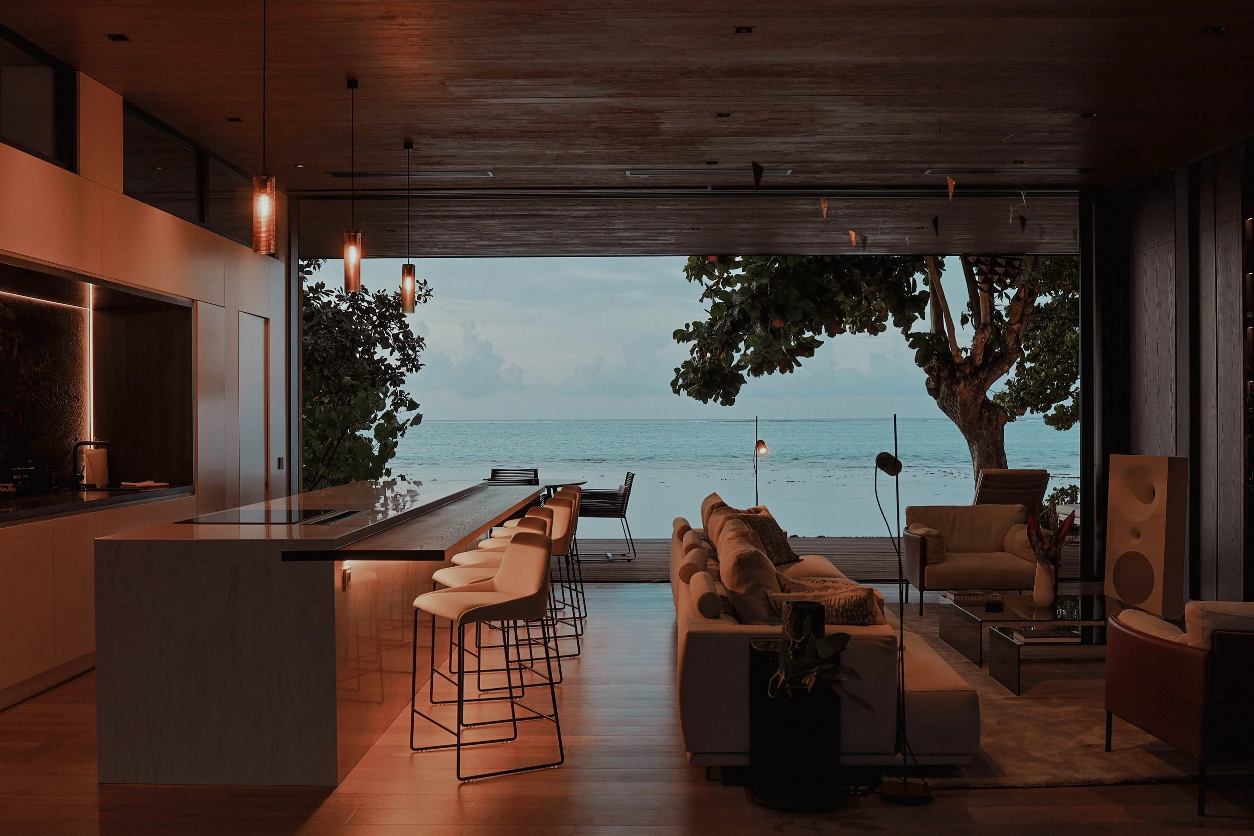 Intérieur d'un salon moderne avec vue sur la mer, comprenant un canapé, des chaises, une table en verre, des lampes design et de grands fenêtres donnant sur la plage et un arbre
