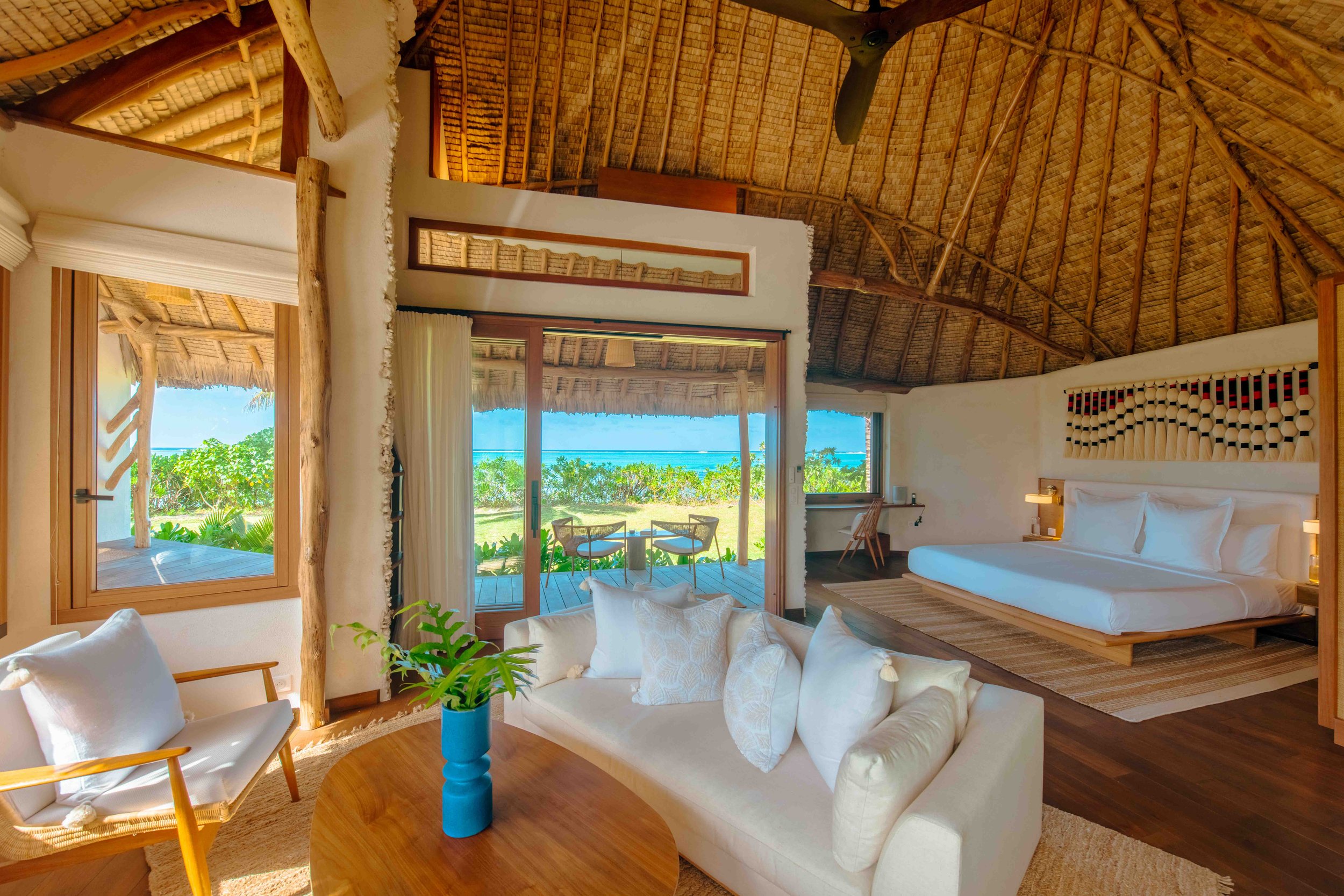Une chambre d'hôtel avec un lit king-size, une décoration tropicale, un salon avec canapé, un bureau, et des portes vitrées donnant sur une terrasse avec vue sur la plage et la mer.