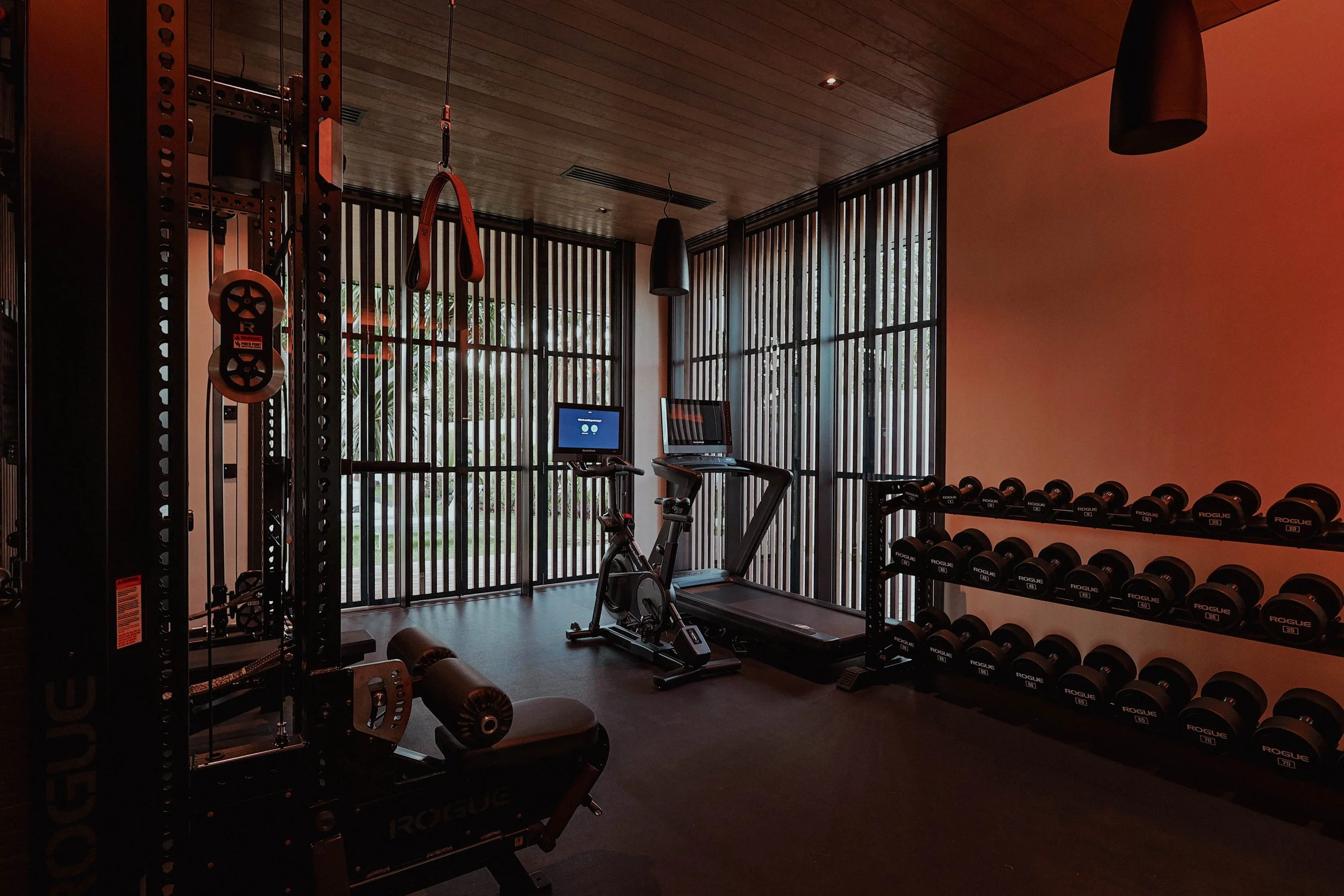 Salle de sport avec tapis de course, vélo d'appartement et rangée de haltères, grandes fenêtres avec volets en bois.