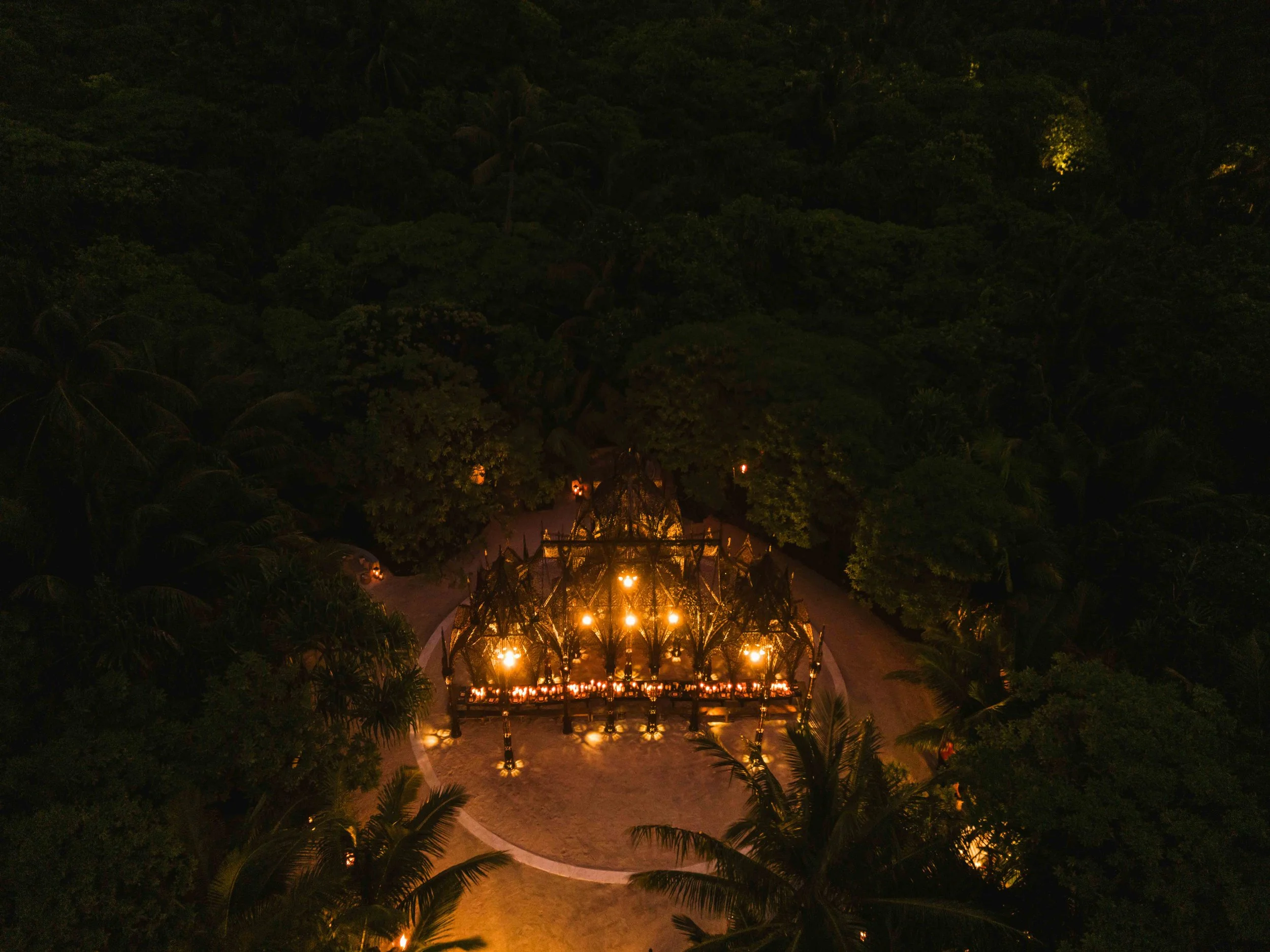Une cérémonie nocturne dans une forêt tropicale, avec des lanternes suspendues et une structure en bois décorée de lumières chaudes.