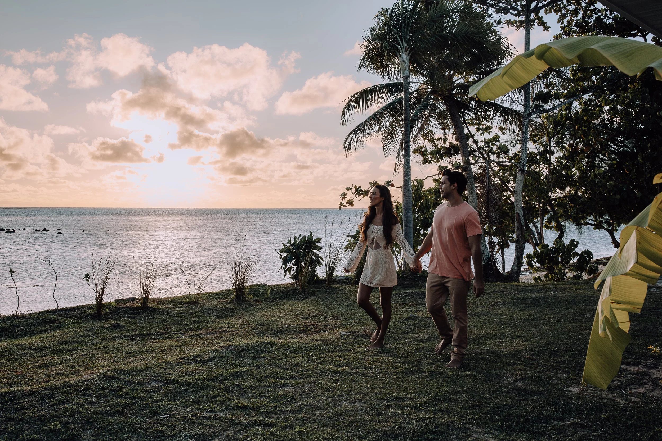 Un couple marche main dans la main sur une pelouse près de la mer au coucher du soleil, entouré d'arbres tropicals.