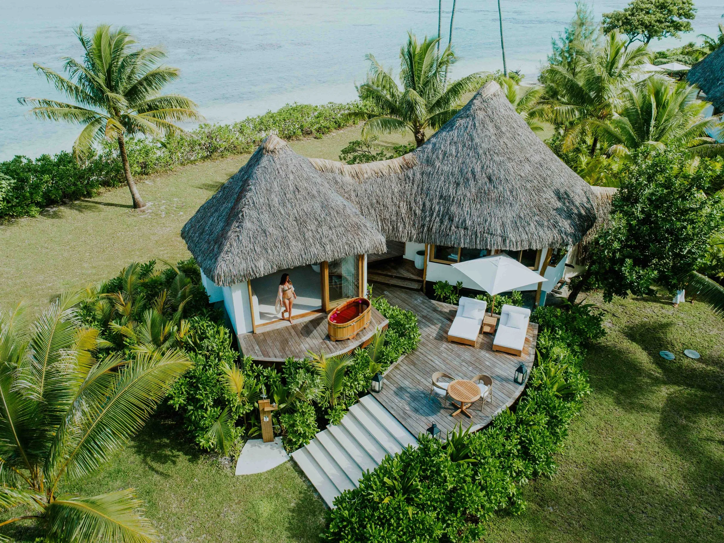 Villa de style tropical avec toits en chaume, entourée de palmiers, située en bord de mer avec une terrasse en bois, meubles de mobilier de jardin, un parasol et une personne debout à l'entrée.