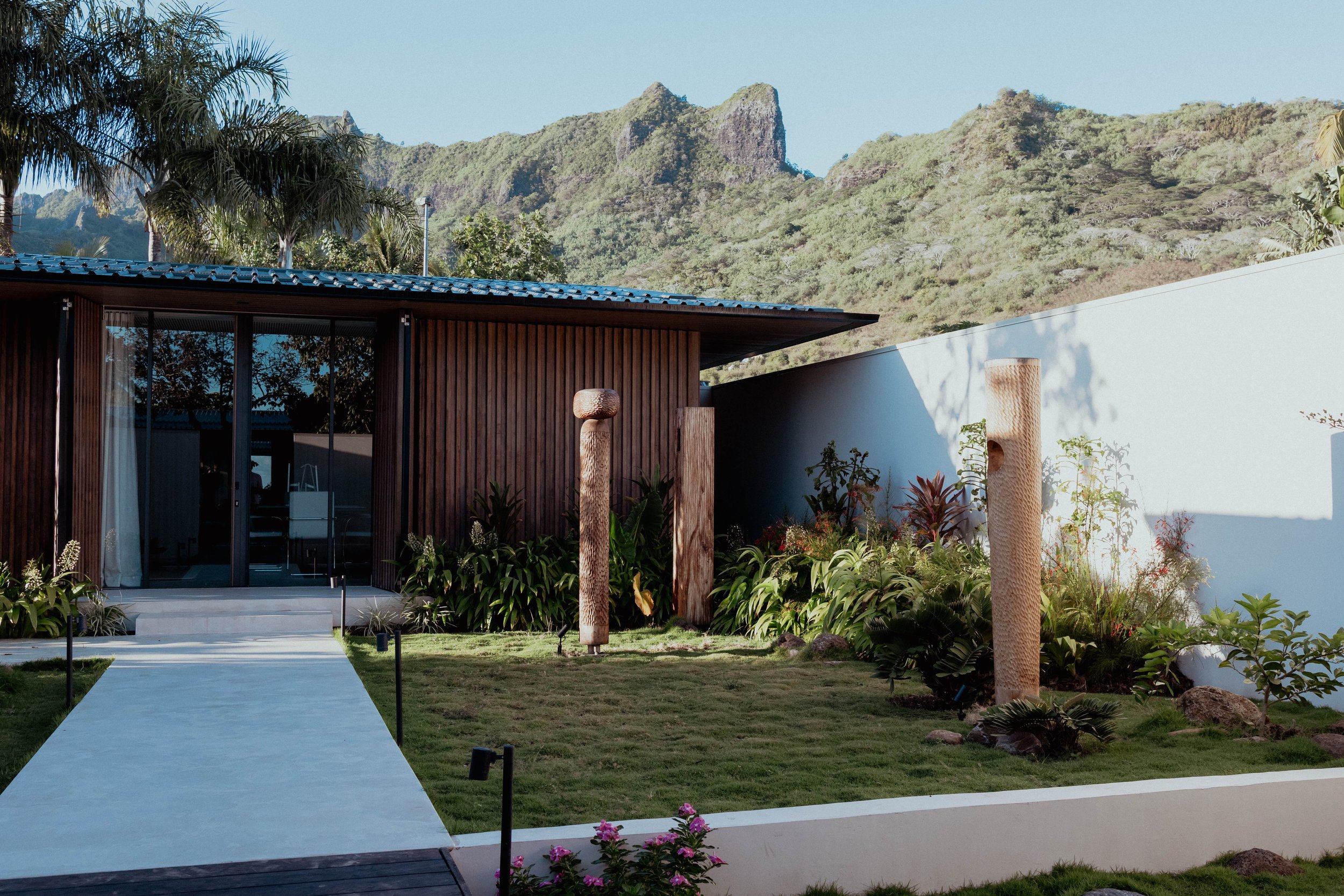Maison moderne avec façade en bois, jardin avec sculptures en bois et plantes, montagnes en arrière-plan, sous un ciel clair.