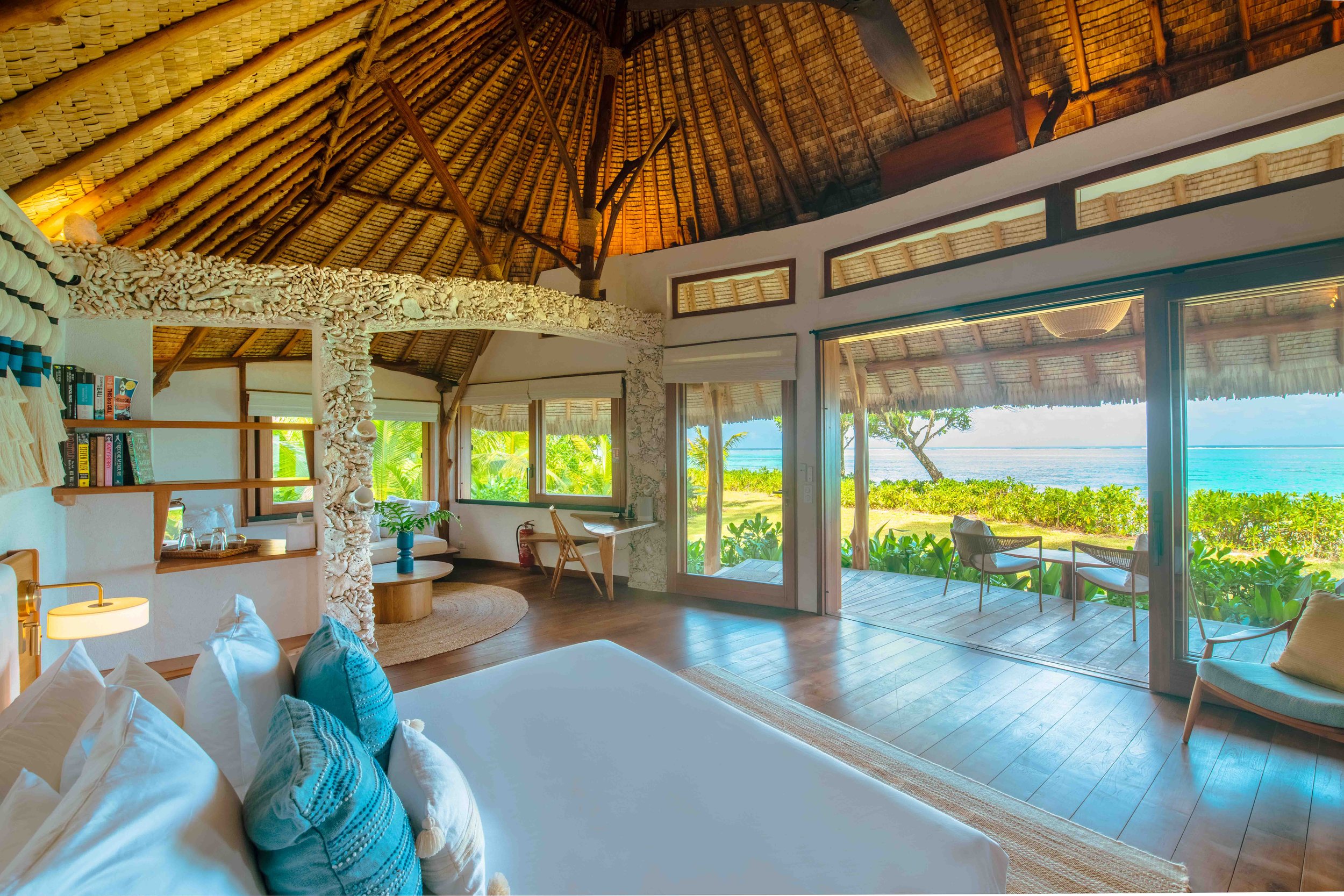 Une chambre spacieuse dans une maison en bord de mer avec un intérieur en bois, un grand lit blanc avec des coussins bleus, et une vue panoramique sur la plage avec des chaises extérieures sur une terrasse en bois.