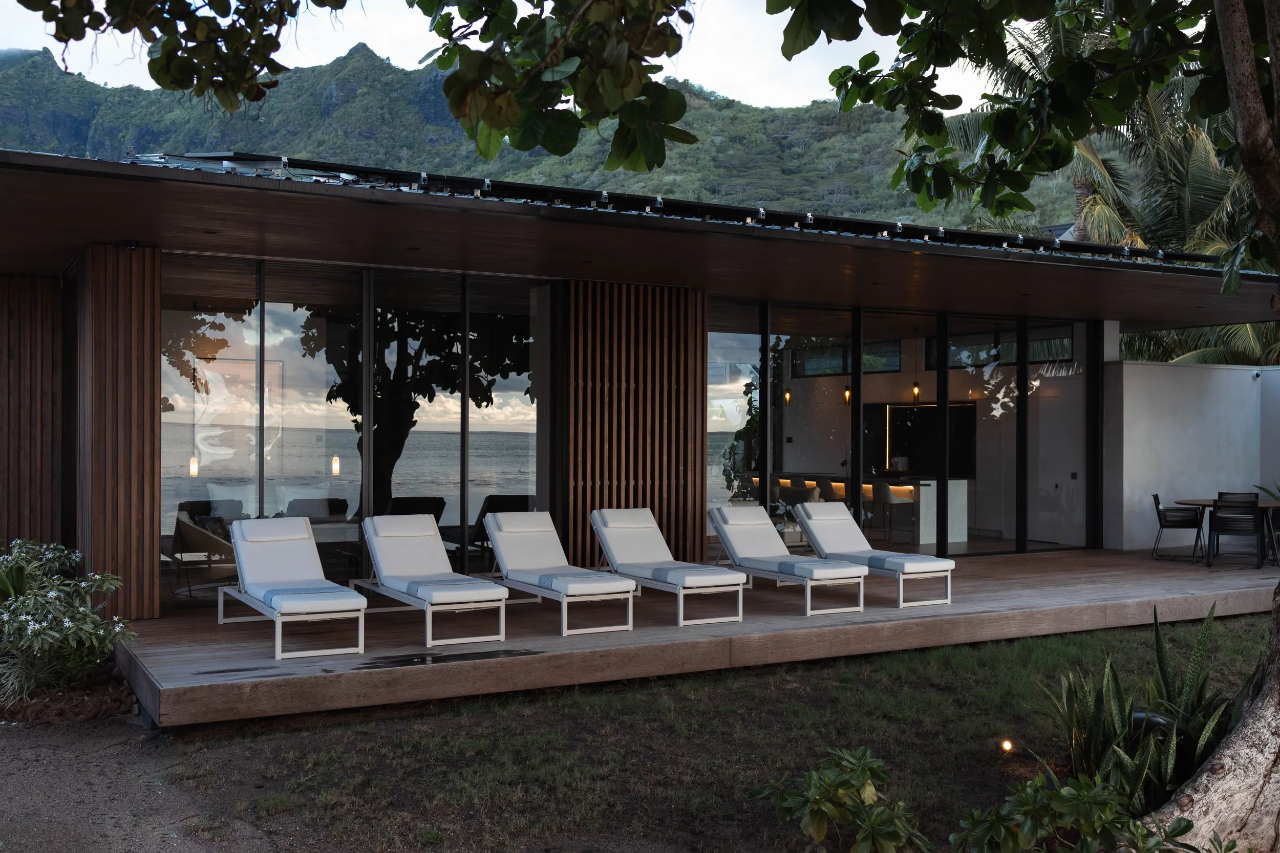 Terrasse en bois avec des transats blancs face à la mer, derrière des fenêtres en verre une maison moderne en bois et en béton avec vue sur la mer et la montagne.