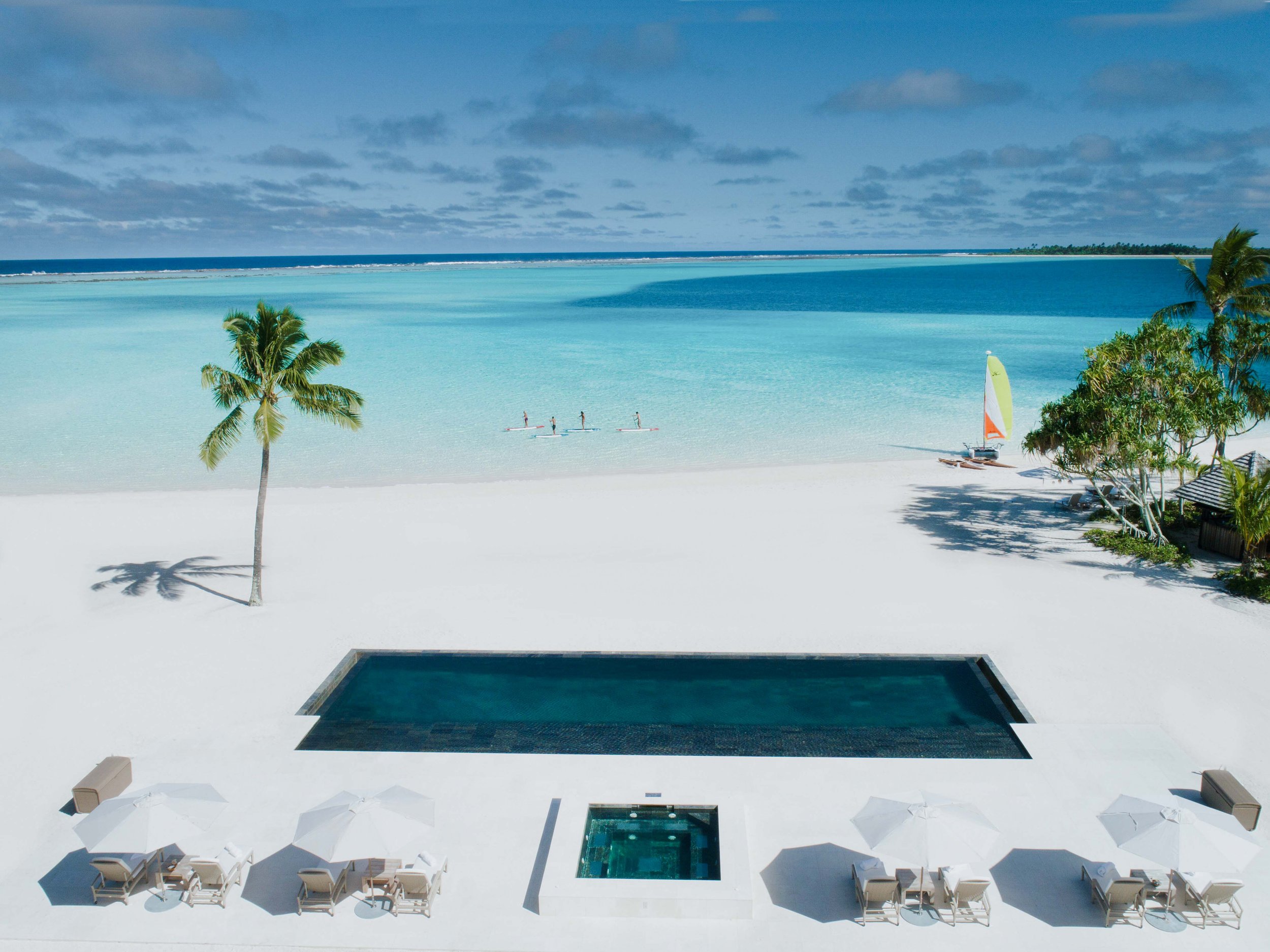 Piscine privée face à une plage de sable blanc, avec chaise-longues et parasols. La mer turquoise avec des sportifs en paddle. Palmiers et végétation luxuriante.