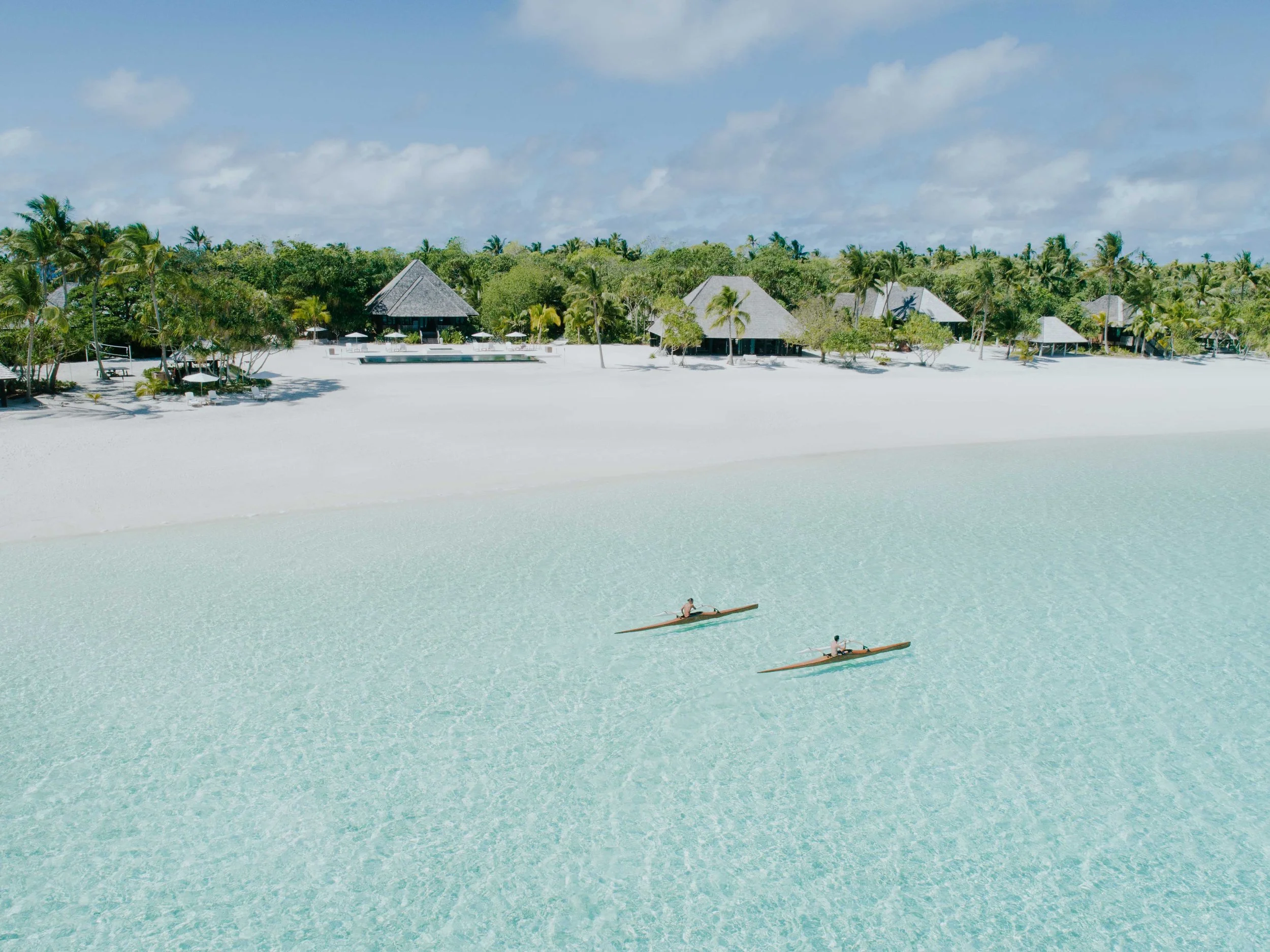 Plage de sable blanc avec deux personnes paddelant en kayak dans une eau turquoise claire, bordée de végétation tropicale et de maisons de vacances en bois avec des toits en chaume.