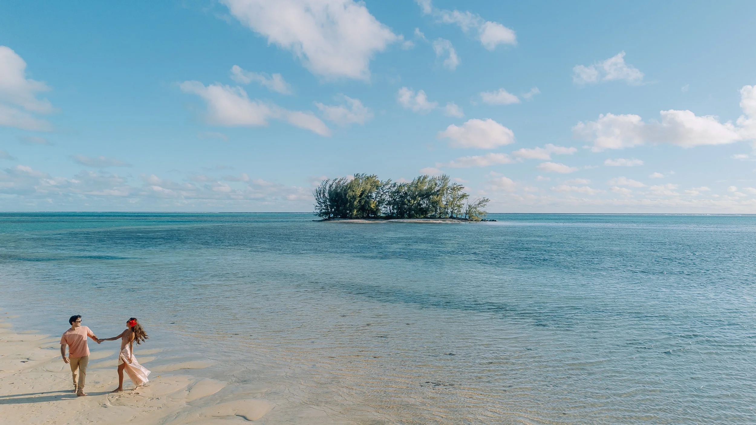 Un couple marche sur une plage de sable blanc près d'une petite île avec des arbres, sous un ciel bleu parsemé de nuages.