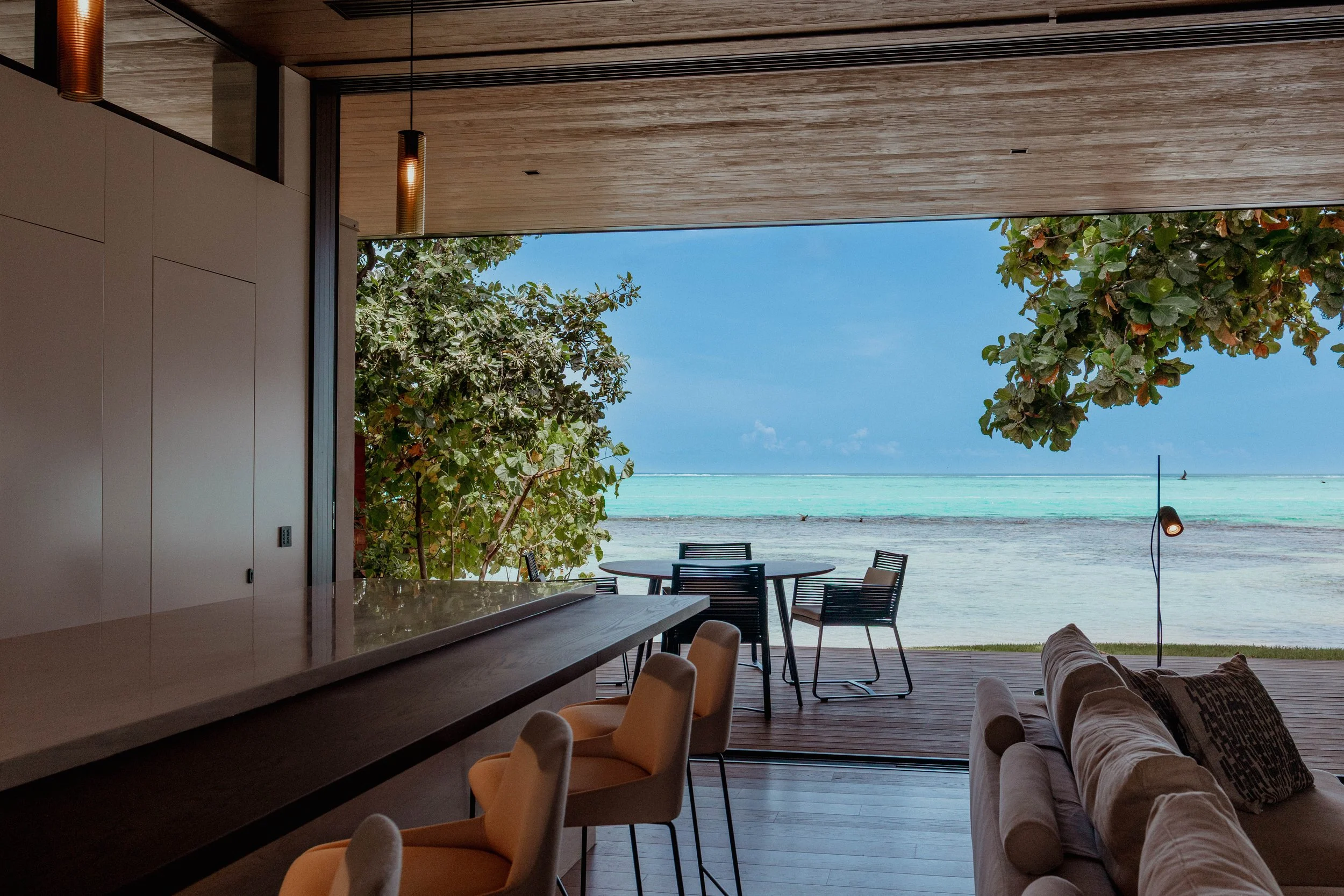Intérieur moderne avec vue sur la plage et la mer turquoise, sièges en velours beige, table à manger, canapé et lampadaire