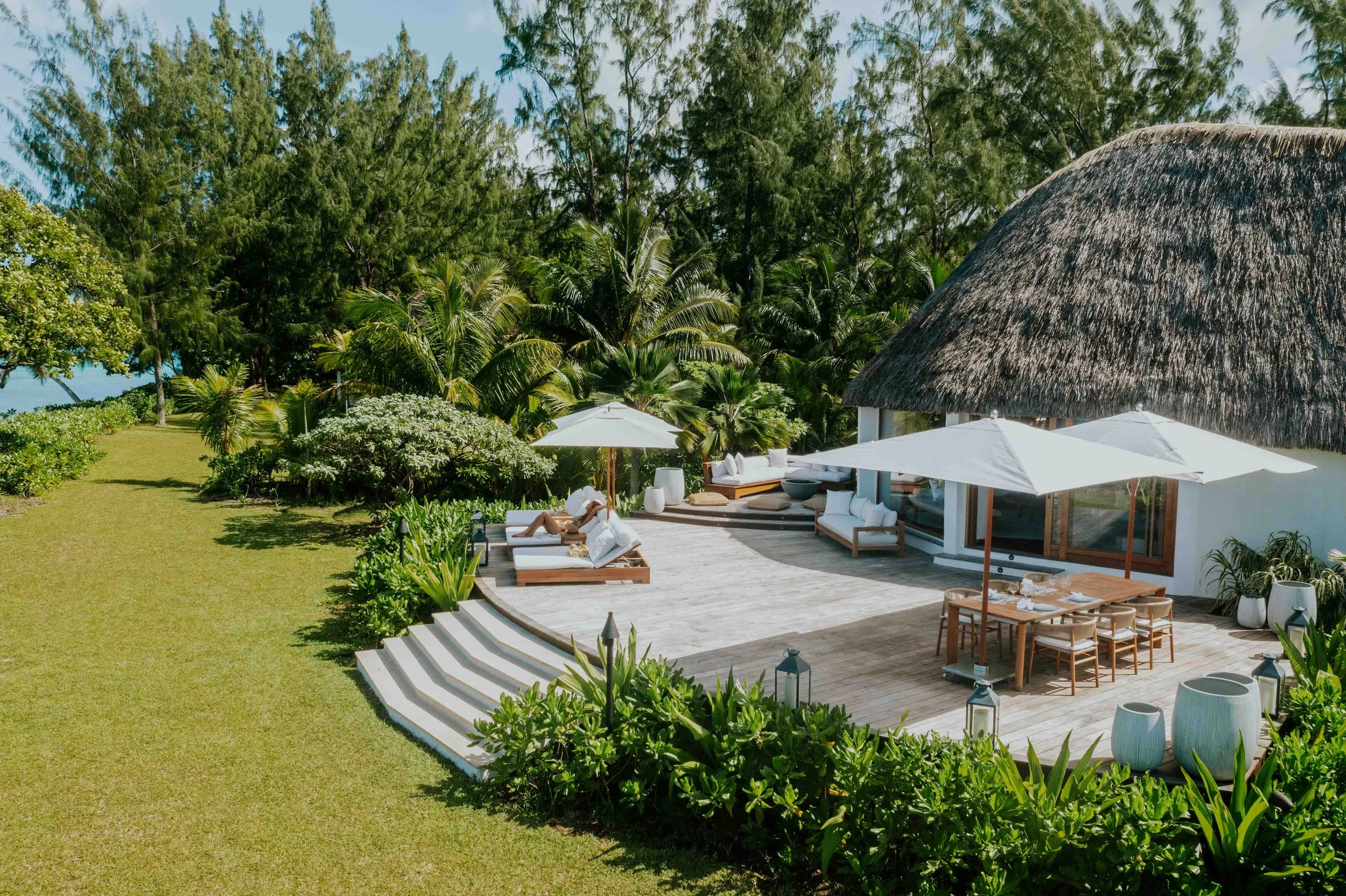 Une maison de style balinais avec un toit en chaume entourée de végétation tropicale, une terrasse en bois avec des parasols blancs, des meubles de jardin et une personne affichant une ambiance relaxante.