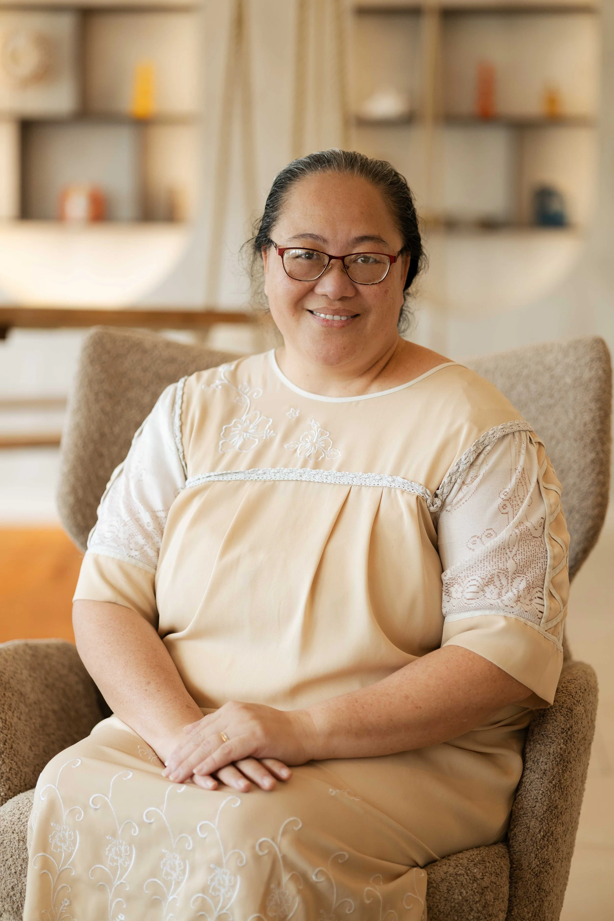 Une femme souriante, portant des lunettes, assise dans un fauteuil en tissu beige, dans un intérieur lumineux avec des étagères en arrière-plan.