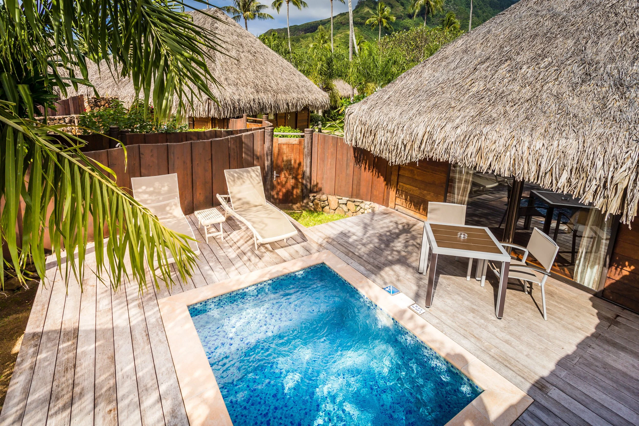 Piscine en contrebas d'une terrasse en bois entourée de cabanes avec toits en chaume, dans un environnement tropical avec des palmiers et des montagnes en arrière-plan.