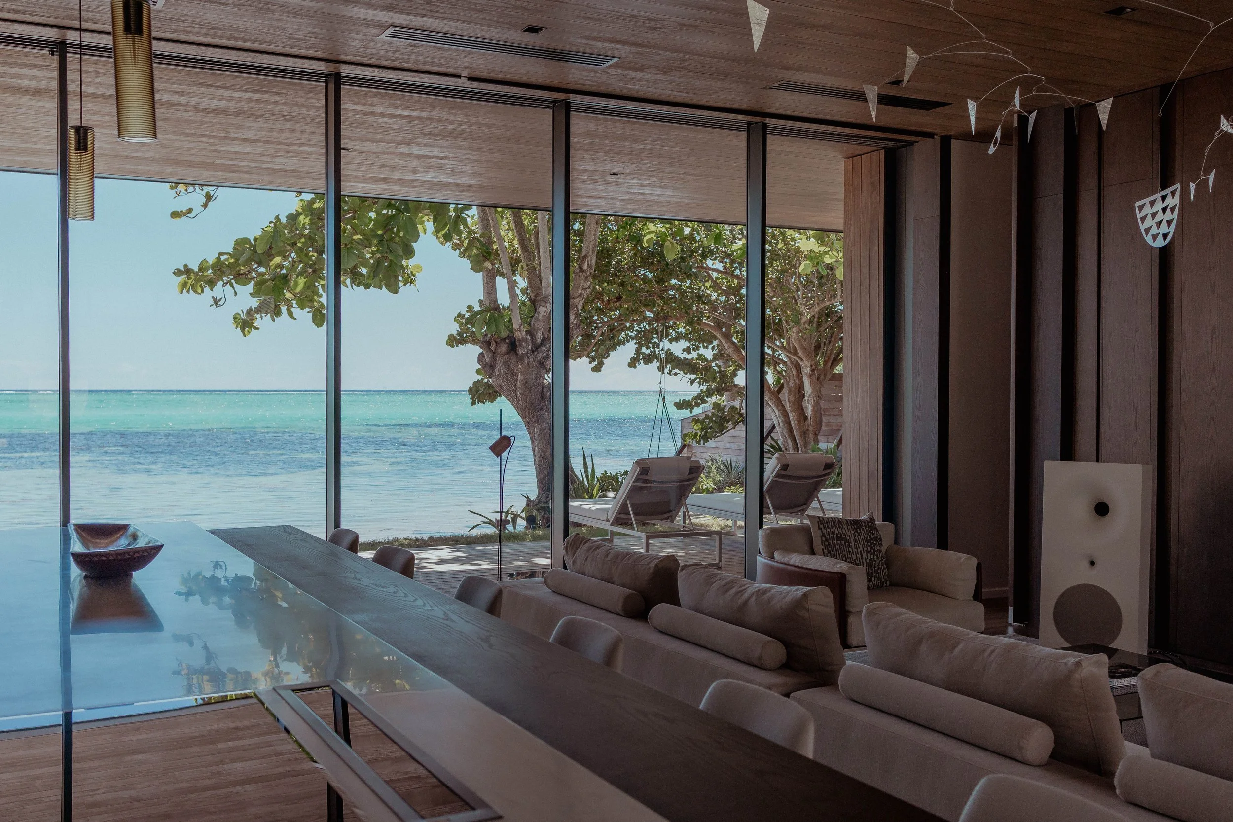 Salon avec vue sur la plage, comprenant des canapés, une table en verre, des chaises, des fauteuils extensions extérieures, des arbres et la mer par la vitre.