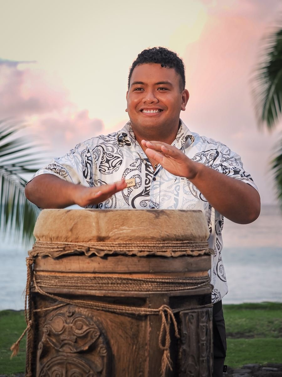 Homme souriant jouant un tambour dans un lieu extérieur avec des palmiers et un coucher de soleil.