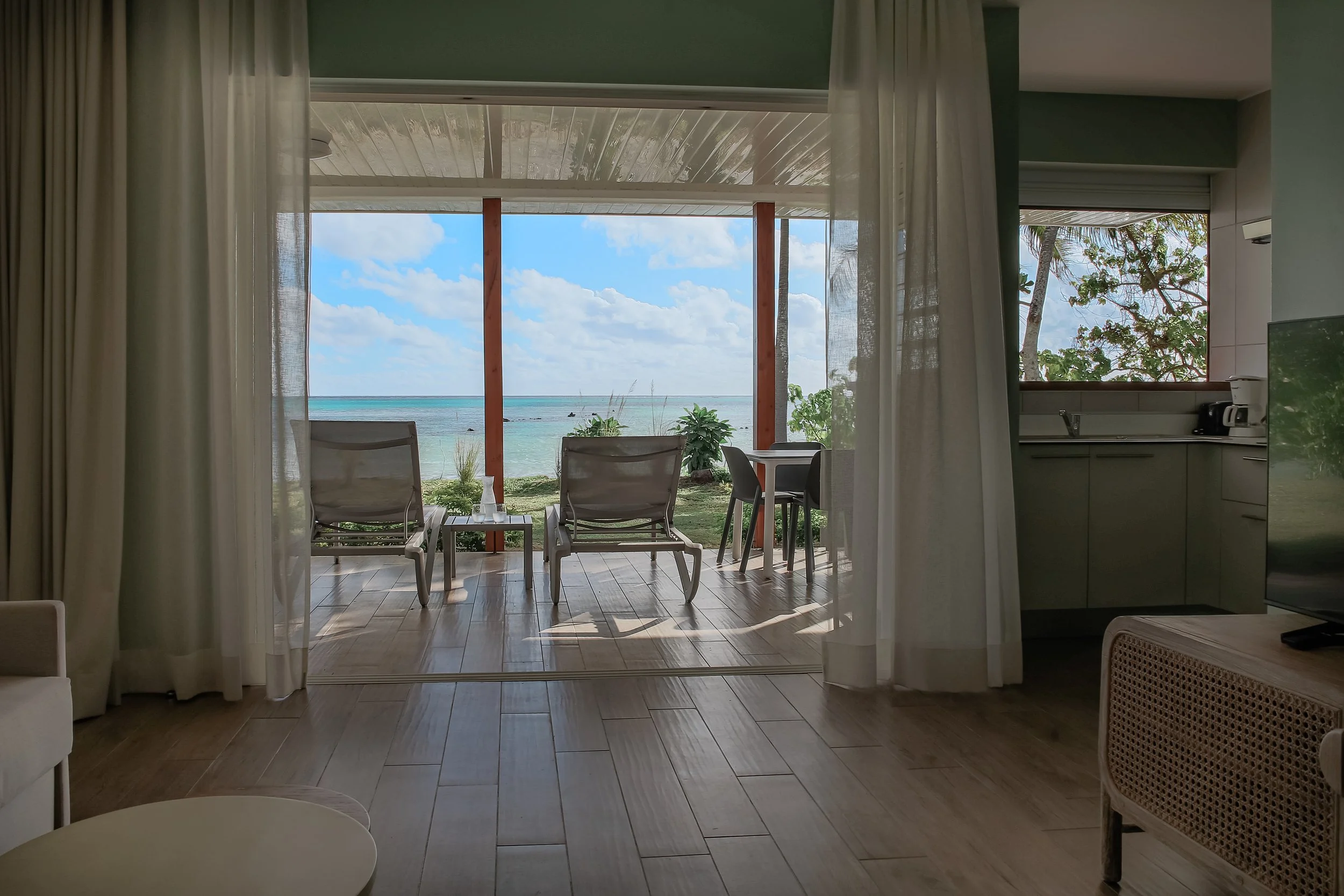 Vue depuis l'intérieur d’un séjour avec porte coulissante ouverte sur une terrasse donnant sur la mer, avec deux chaises longues et un petit tableau, sous un ciel bleu avec quelques nuages.