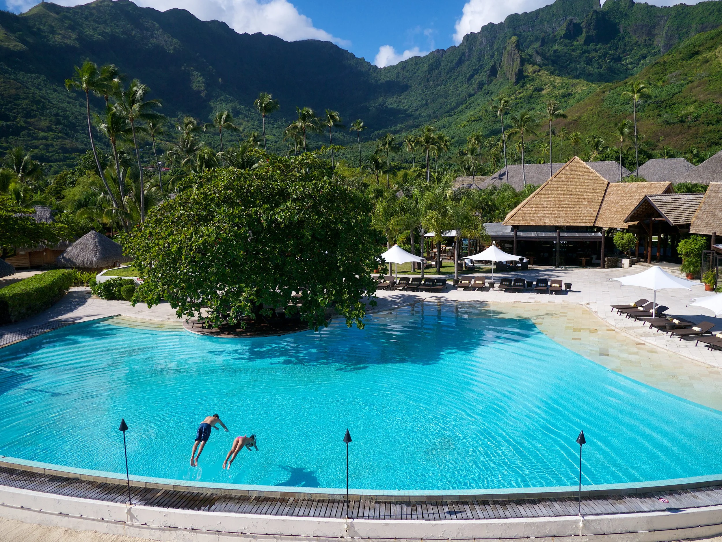 Deux enfants en maillot de bain nageant dans une piscine située dans un complexe hôtelier en milieu tropical, avec des palmiers, des montagnes verdoyantes en arrière-plan, et des cabanes en bois avec des toits en chaume.