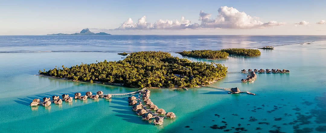 Vue aérienne d'une île tropicale avec des bungalows sur pilotis reliés par des ponts, entourée d'eau turquoise et de nuages en arrière-plan.