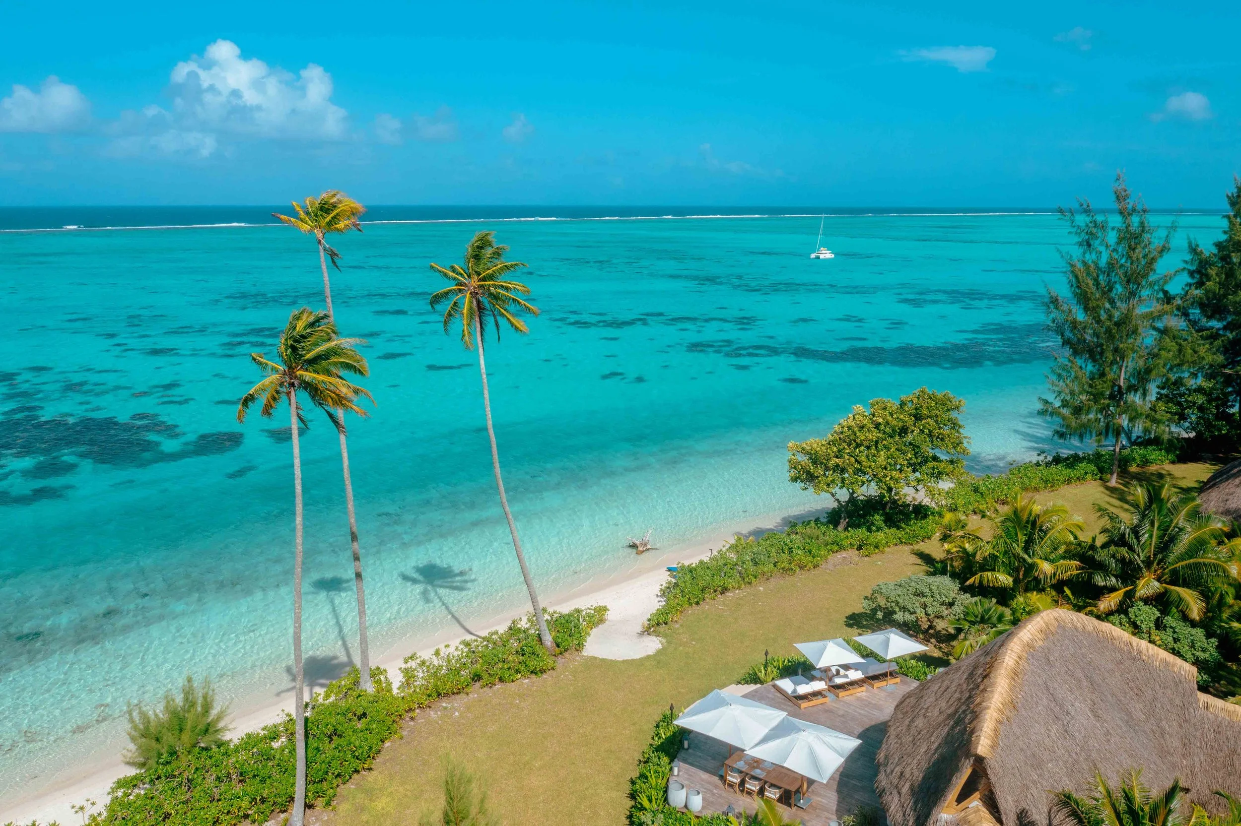 Plage tropicale avec sable blanc, palmiers, et cabanes avec parasols blancs, vue sur la mer turquoise et le ciel bleu avec quelques nuages.