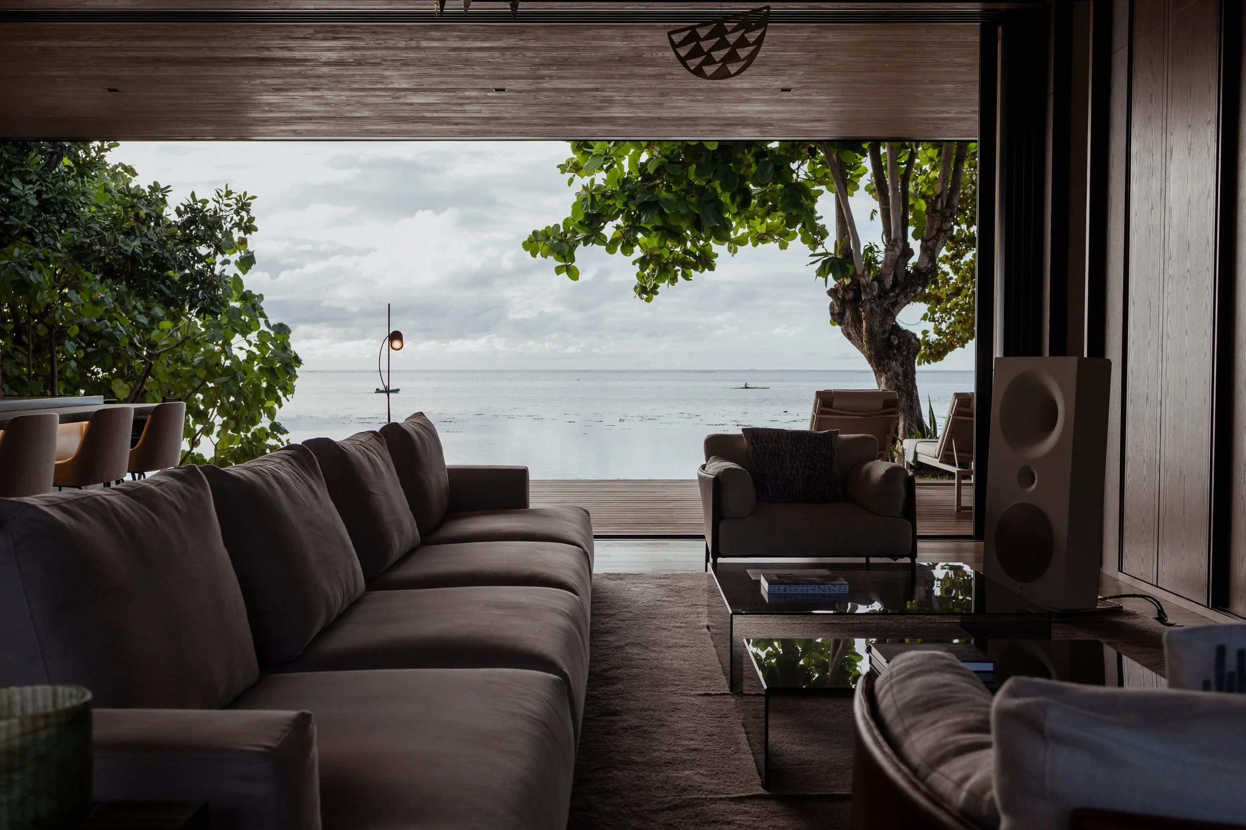 Intérieur d'un salon moderne avec vue sur la mer, canapé gris, une table basse en verre, un fauteuil, un haut-parleur blanc, et des chaises avec vue sur l'eau et un arbre outside.