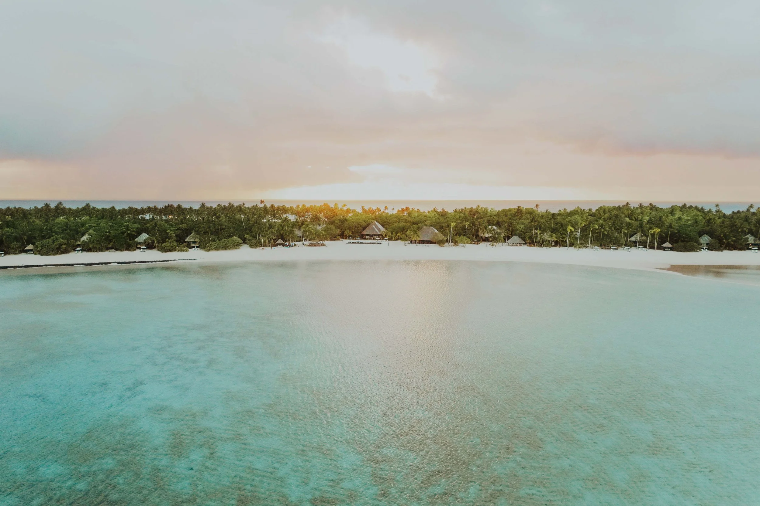 Plage de sable blanc avec des cabanes en bois, eau turquoise calme et forêt dense en arrière-plan, coucher de soleil