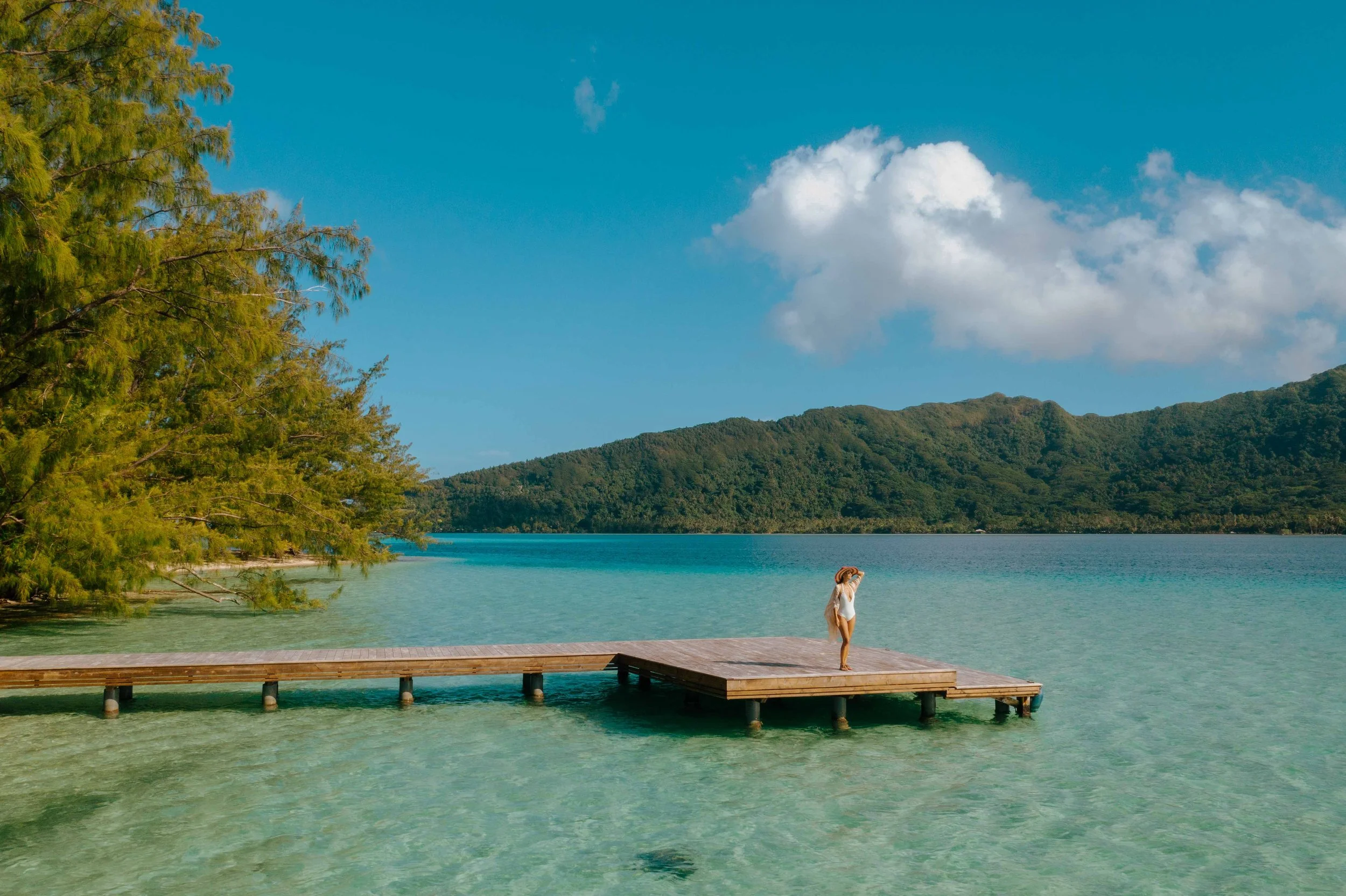 Une femme sur une jetée en bois au bord d'un lac turquoise entouré de montagnes verdoyantes, sous un ciel bleu avec quelques nuages.