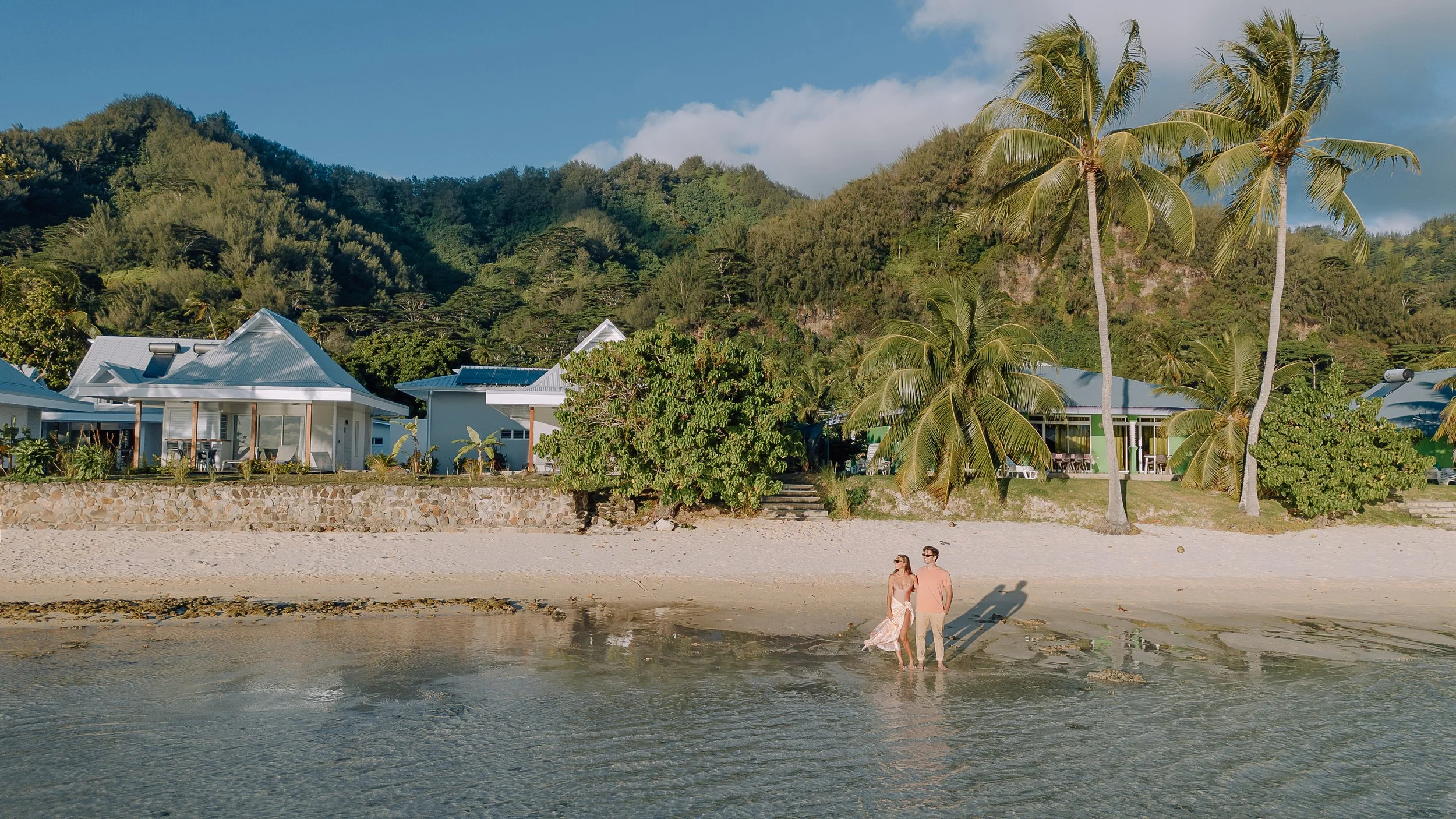Un couple marche sur la plage devant des maisons blanches avec un toit en métal, entourées de palmiers et de verdure, avec une montagne en arrière-plan et un ciel bleu.