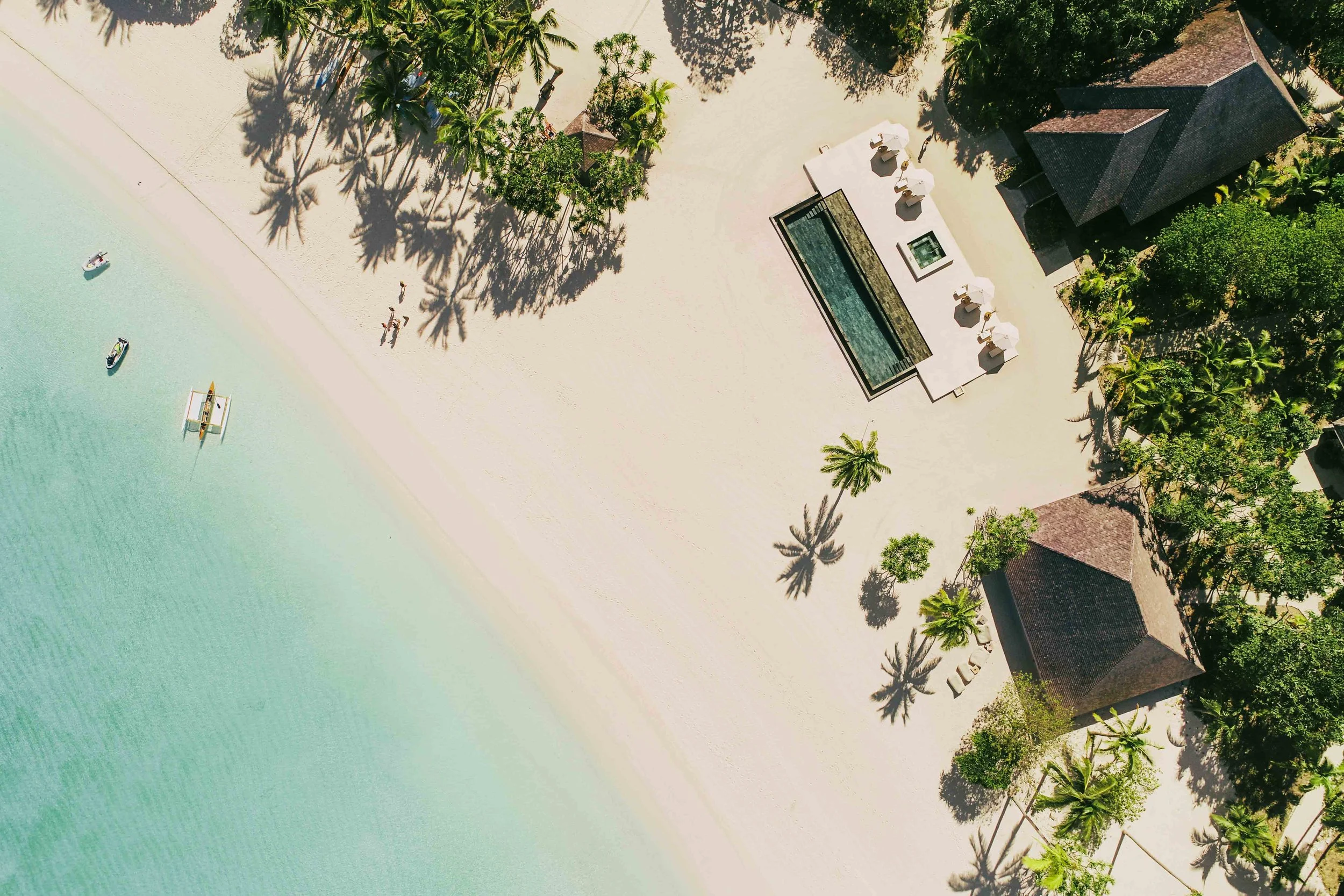 Vue aérienne d'une plage de sable blanc avec des parasols, une piscine, des villas, et quelques palmiers à l'ombre.