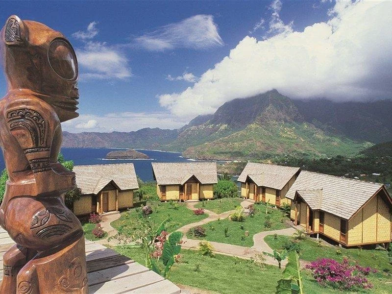 Vue panoramique de cottages en bois entourés de jardins verdoyants avec des fleurs, face à une grande étendue d'eau et des montagnes en arrière-plan, avec un sky bleu et quelques nuages, et une sculpture en bois sur le côté gauche.