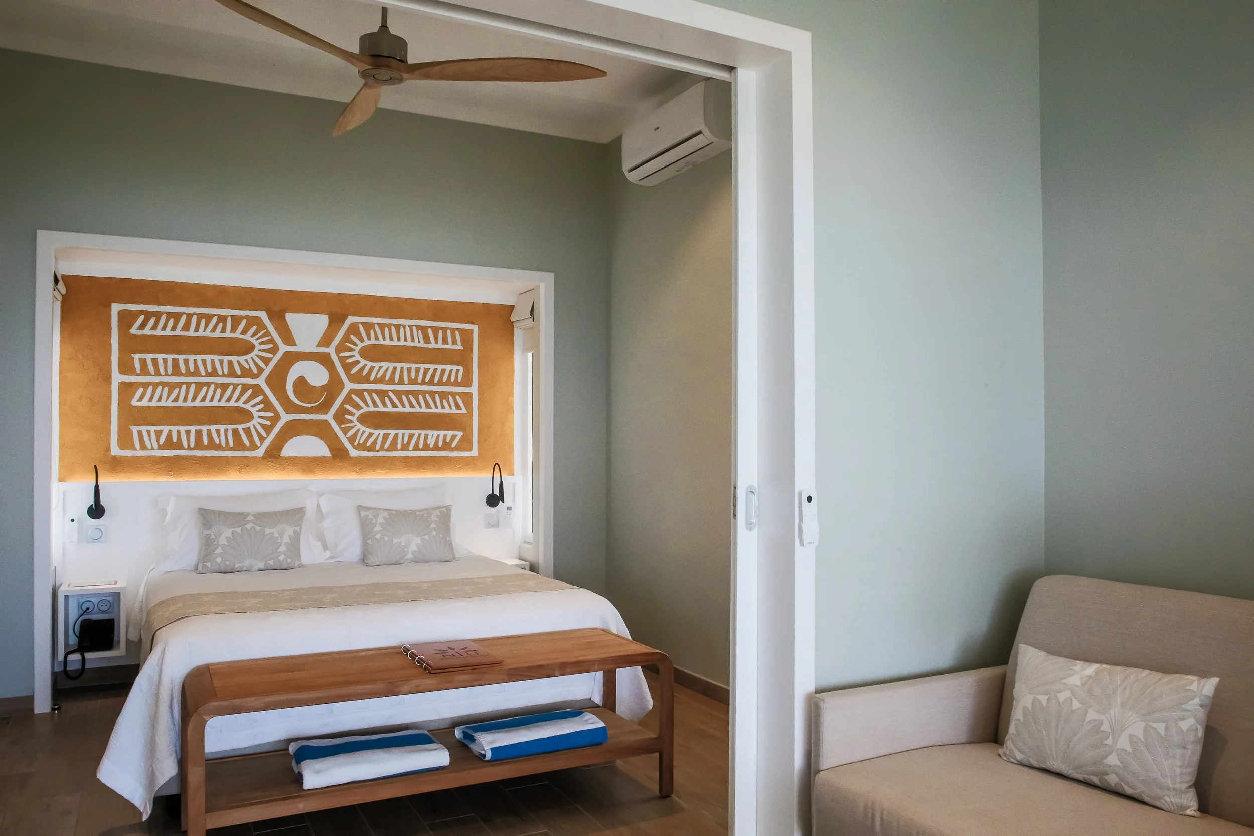 Chambre d'hôtel moderne avec un lit blanc, une tête de lit ornée d'une œuvre d'art murale en bois, et un coin salon avec un canapé beige et un coussin.