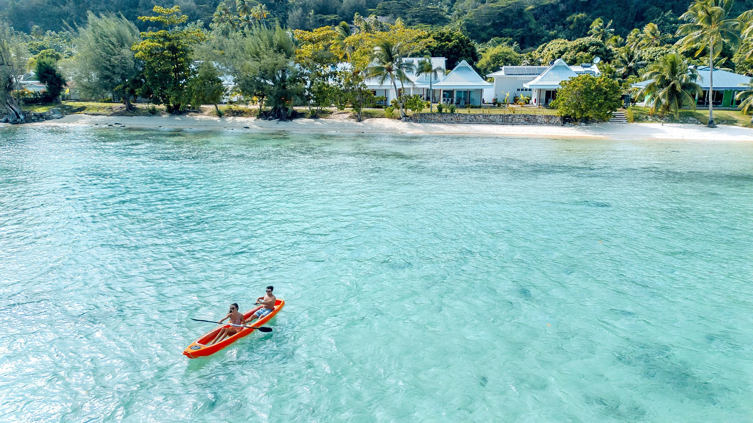 Deux personnes en kayak orange sur une eau turquoise calme, avec une plage bordée d'arbres et des maisons en arrière-plan.