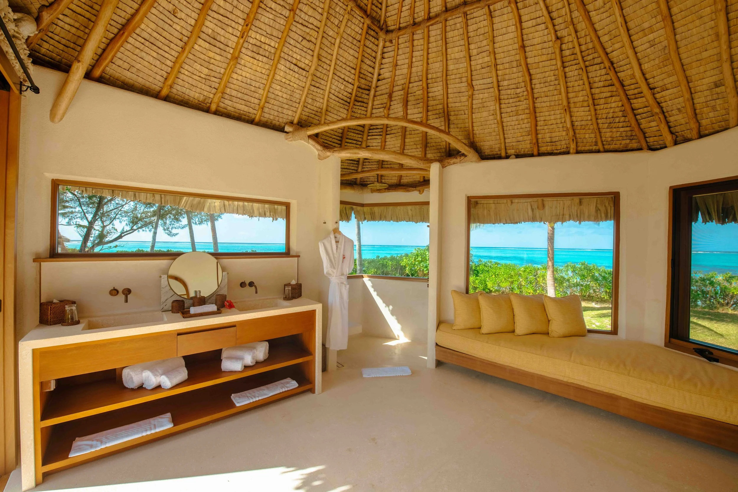 Intérieur d'une cabane de style tropical avec vue sur la plage, comprenant un lavabo avec un miroir, une banquette beige avec des coussins, et de grandes fenêtres offrant une vue sur la mer et les arbres.