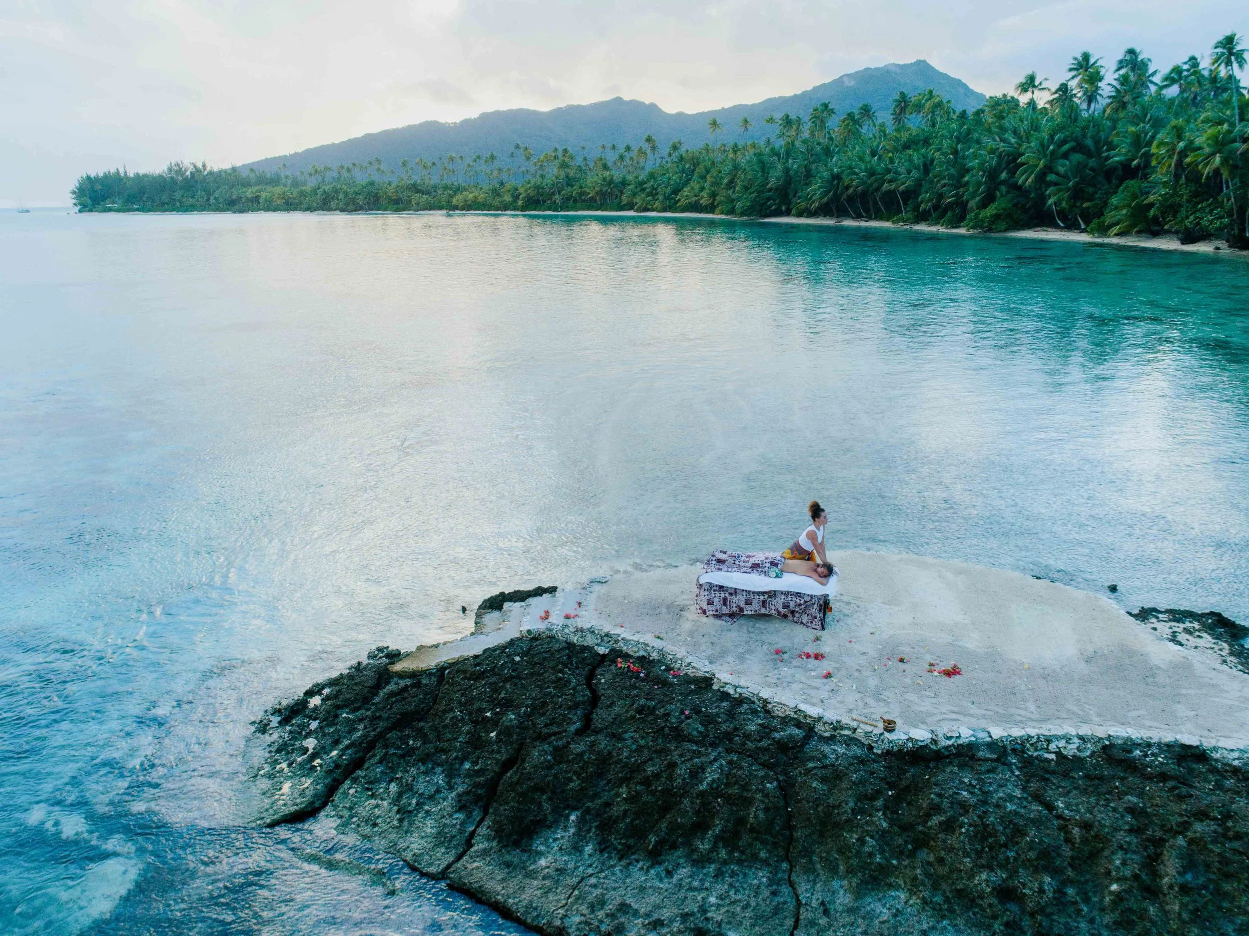 Une femme recevant un massage sur une plateforme en bord de mer avec des palmiers et des montagnes en arrière-plan.