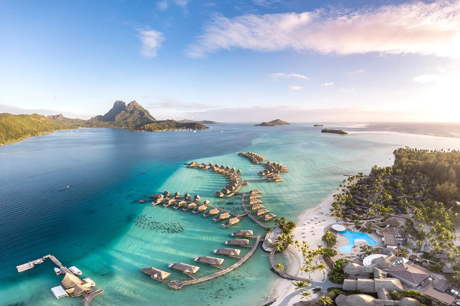 Vue aérienne d'une station balnéaire avec des bungalows sur pilotis en forme de chaîne, une piscine d'hôtel, une plage de sable blanc avec des palmiers, et une mer turquoise à l'île de Bora Bora avec le Mont Otemanu en fond.