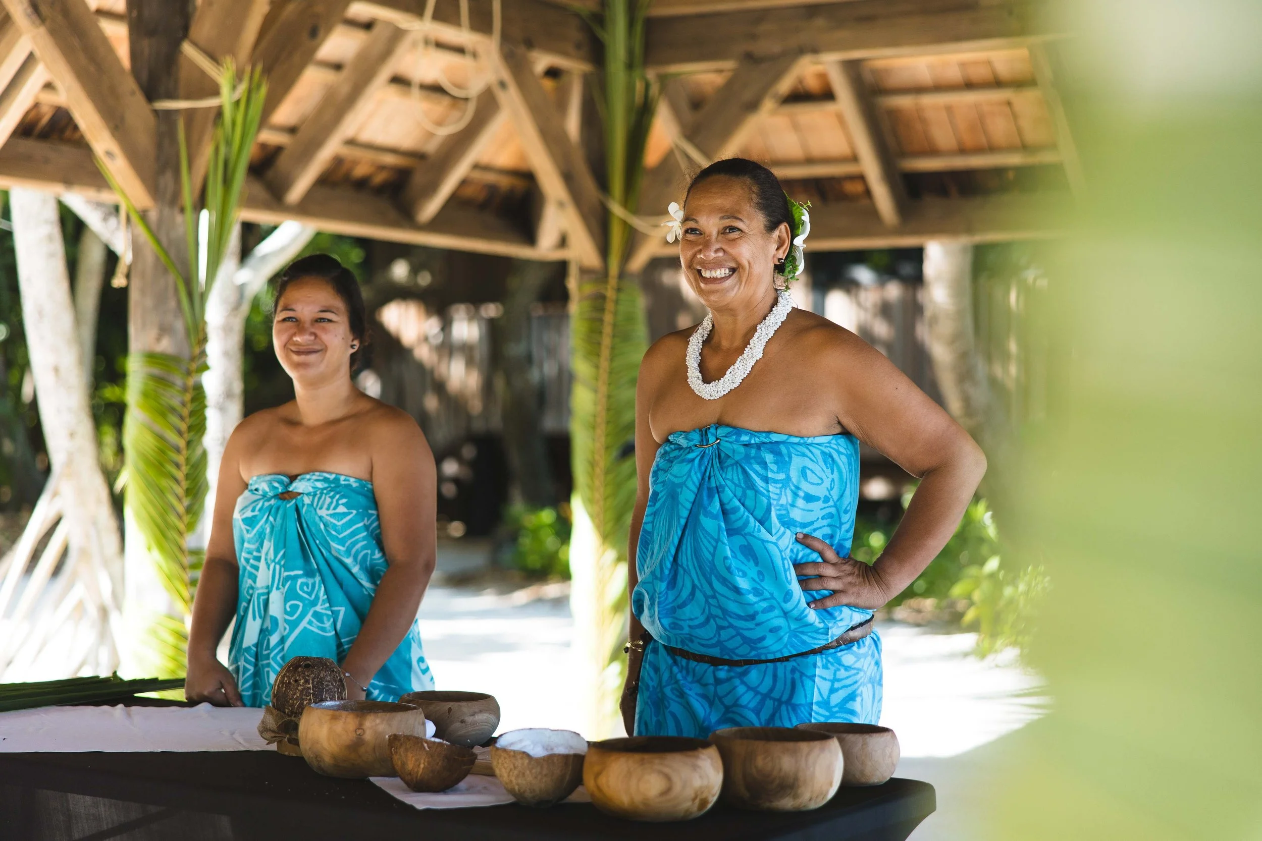 Deux femmes Polynésiennes souriantes, vêtues de wraps bleus traditionnels, se tiennent derrière une table avec des bols en bois contenant des ingrédients culinaires, sous un cabanon en bois avec des feuilles de palmier.