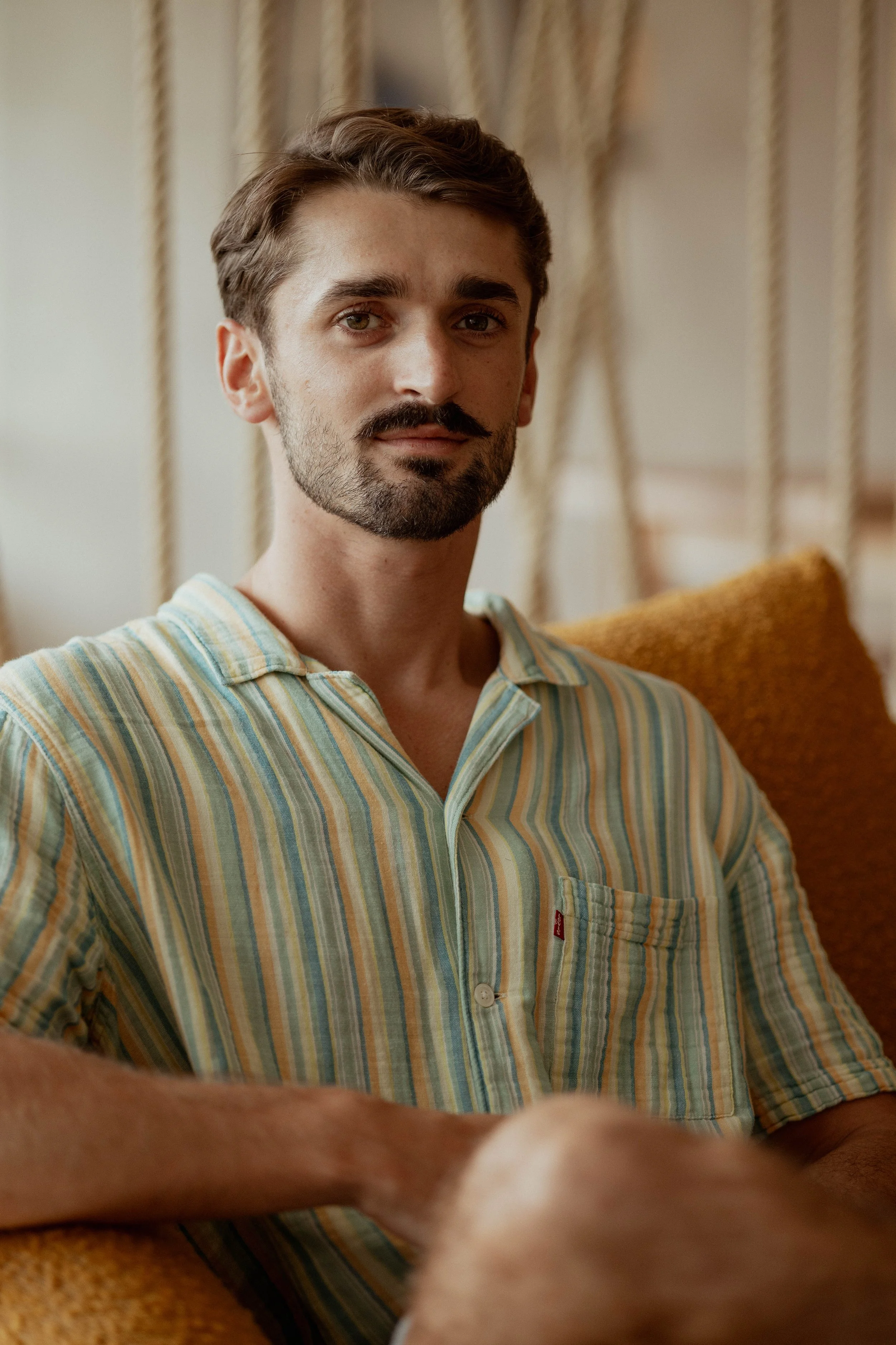 Un homme à barbe, portant une chemise à rayures, assis sur un canapé en tissu orange avec des cordes comme décoration en arrière-plan.