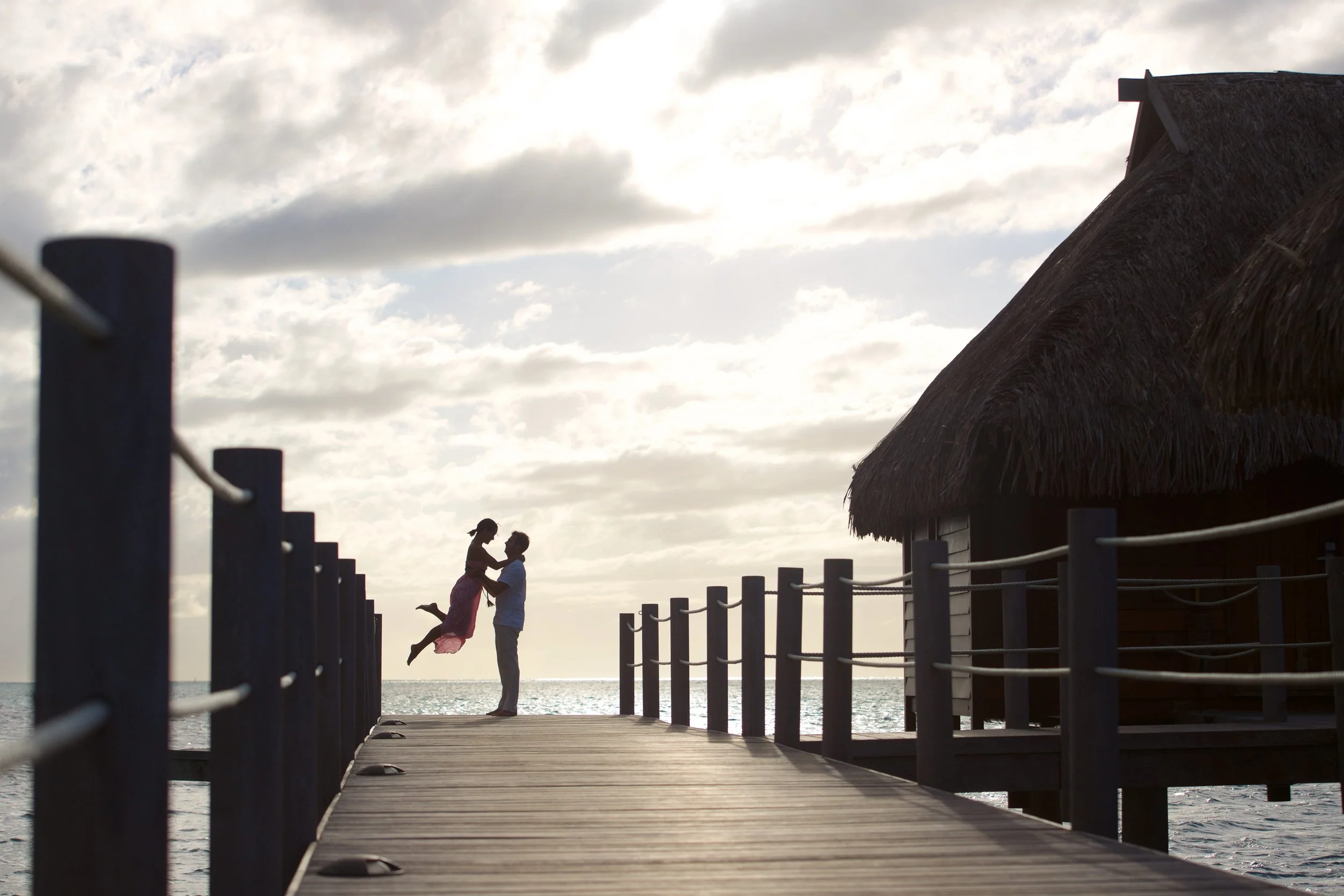 Un homme et une femme sur un pont en bois au bord de la mer, avec un ciel nuageux. La femme est en l'air, tenue par l'homme, tous deux regardant l'un l'autre, avec une cabane en bois à toits de chaume à droite.