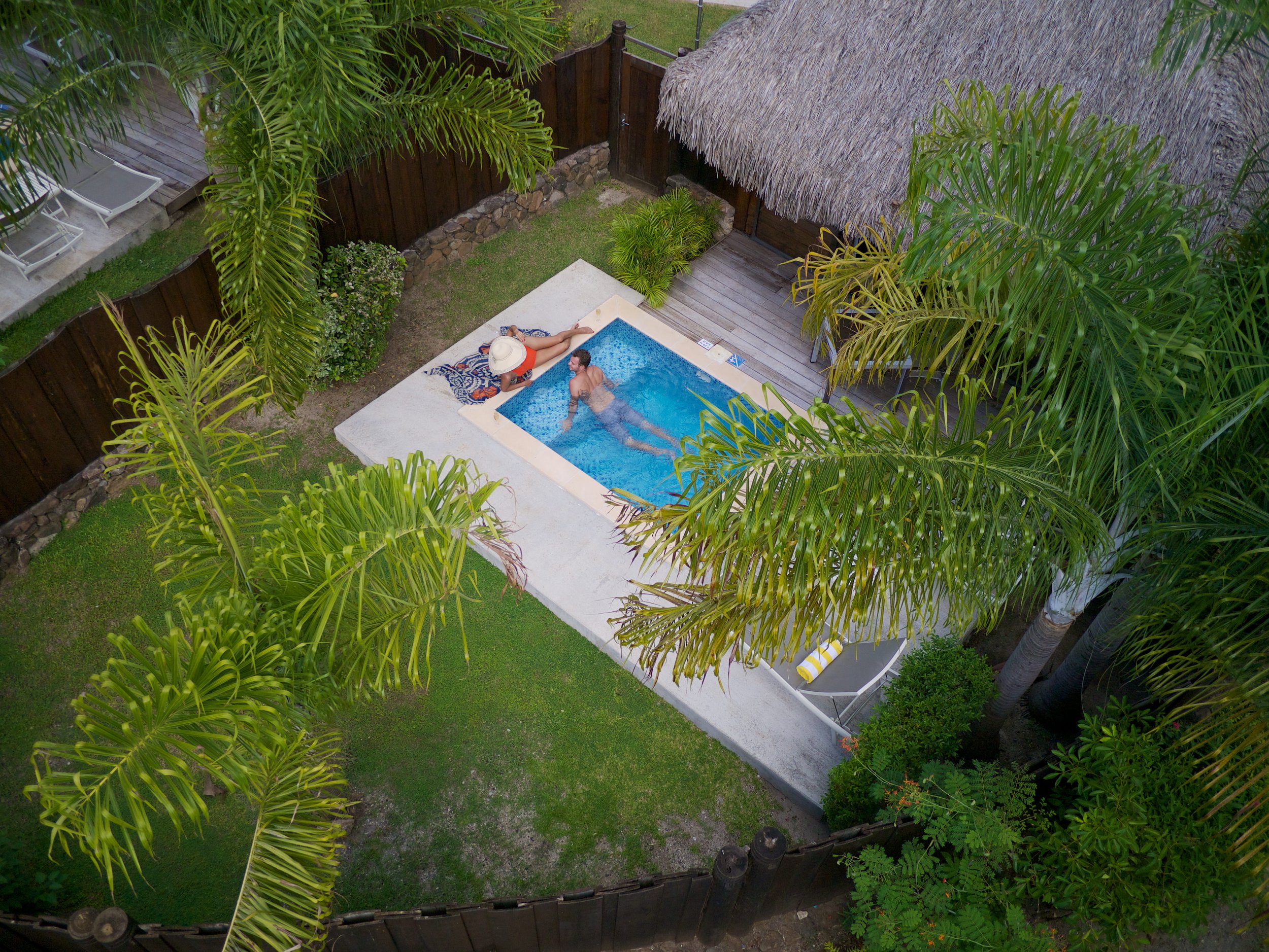 Deux personnes dans une petite piscine dans un jardin tropical entouré de palmiers, avec un parasol, une chaise longue et une cabane en toit de chaume.