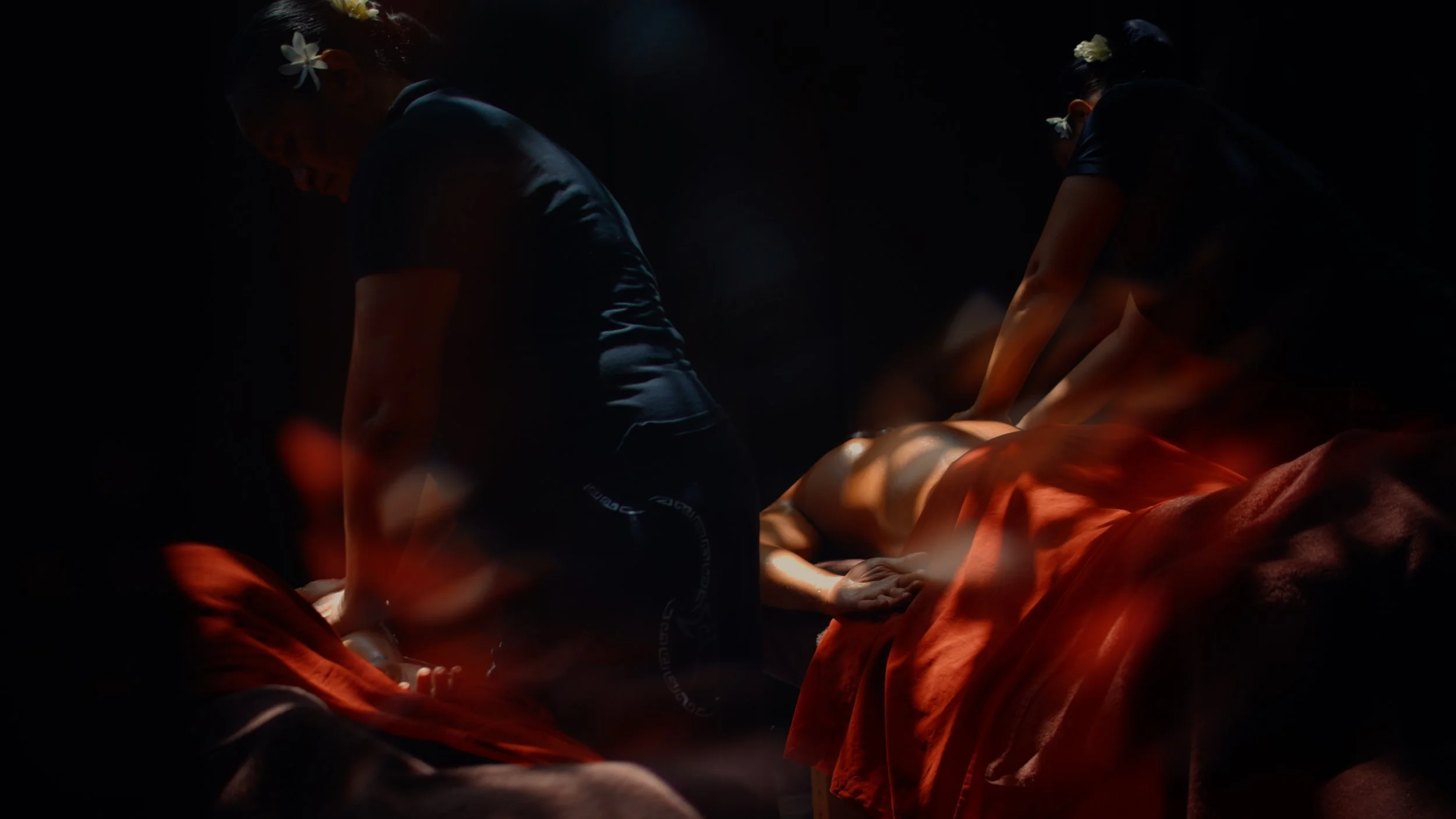 Deux personnes donnent un massage sur un matelas enroulé dans un drap orange, dans une pièce sombre, avec un éclairage doux et une ambiance apaisante.